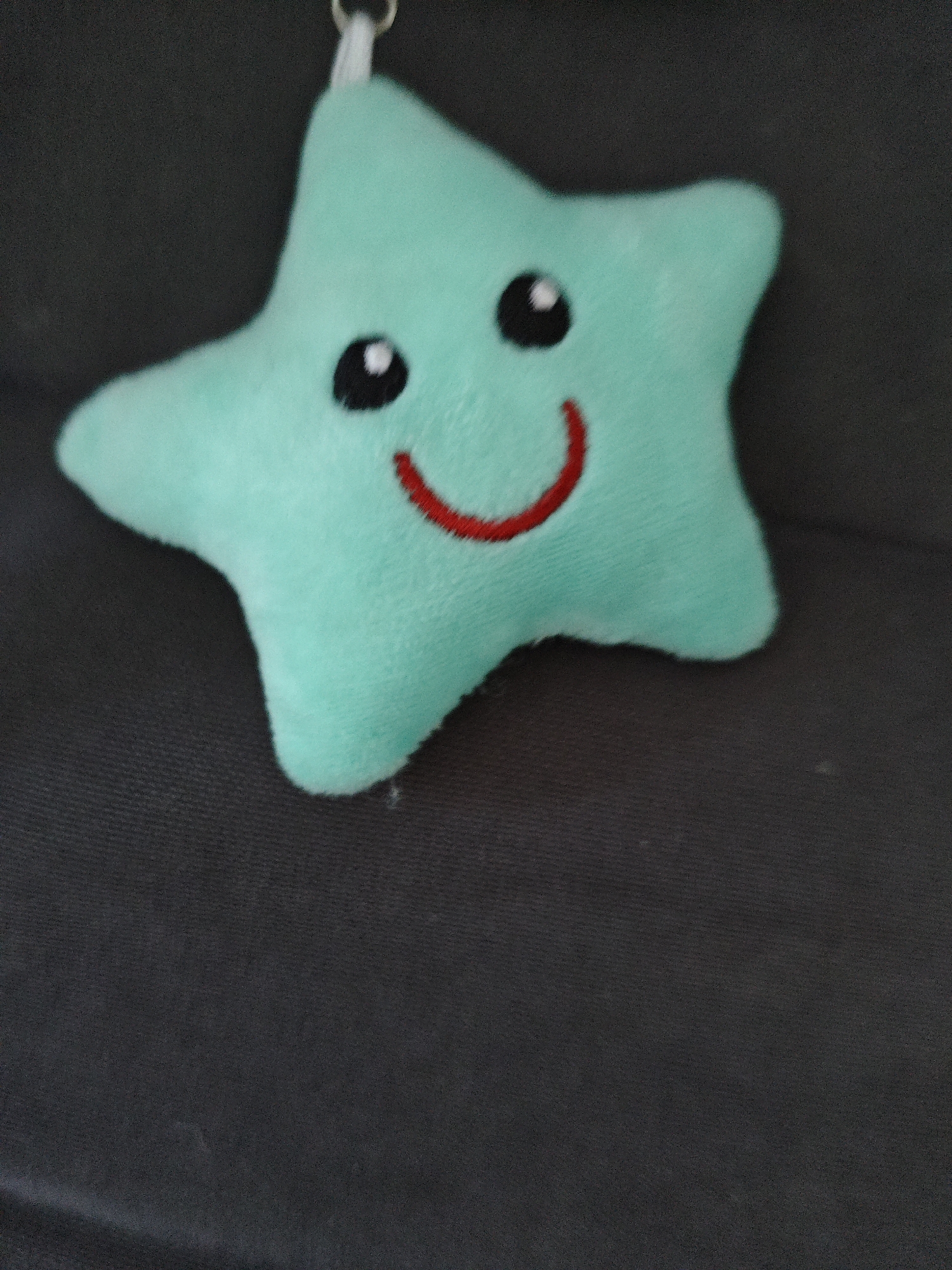 A plush turquoise star-shaped keychain with embroidered black eyes and a red curved mouth is laid flat on a dark gray textured surface. The keychain has a small silver ring attached to one of its points. The lighting suggests an indoor setting. The image does not provide any information or context to determine its location in Alushta, Ukraine.