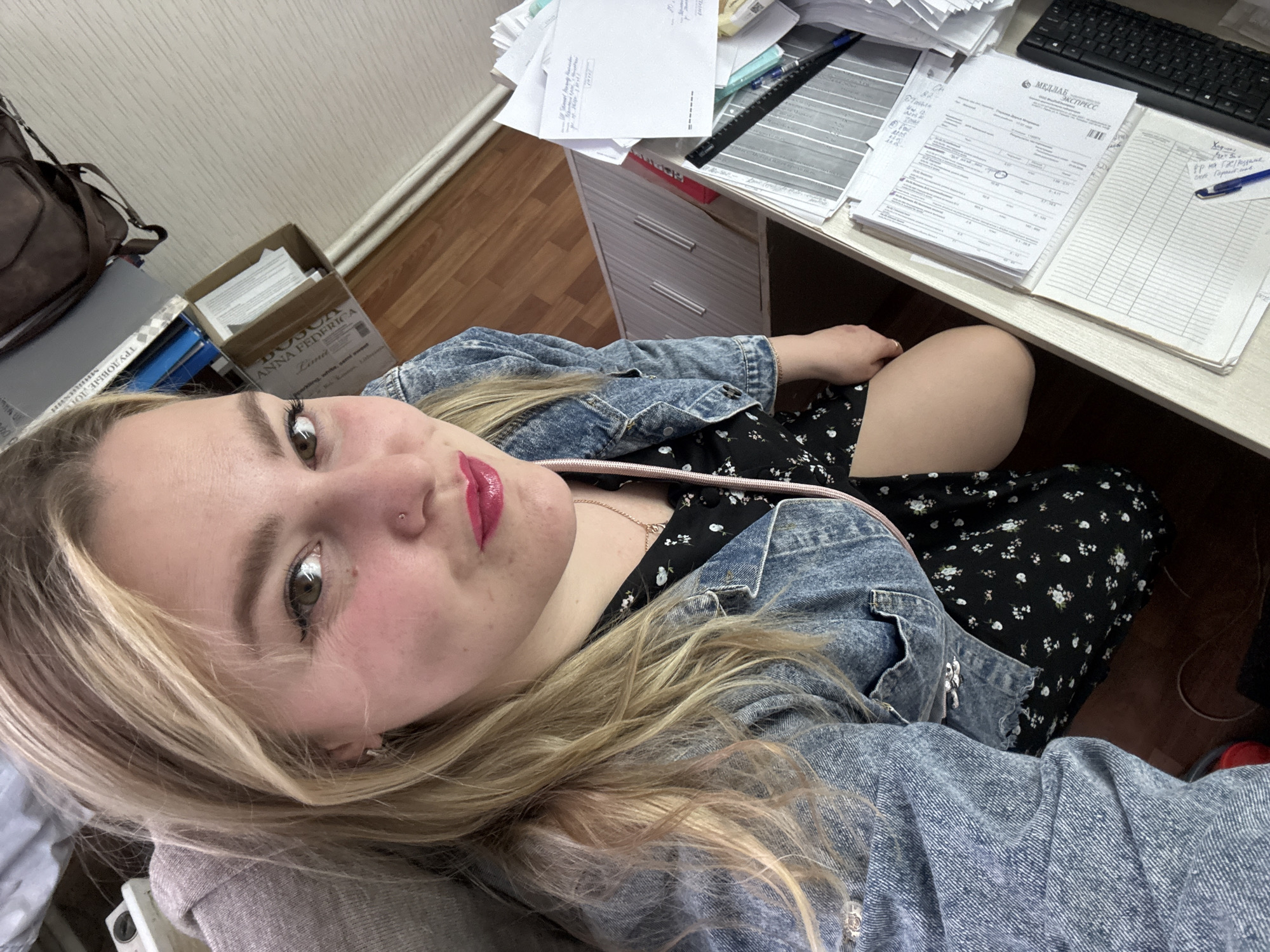 The image shows a fair-skinned young woman with blonde hair, wearing a denim jacket over a black floral dress, seated at a desk. She is looking directly at the camera, posing for a selfie. There is a document on the desk labeled "МЕДЛАБ КСПРЕСС" and "ANNA FEDERICA Lima" indicating the location in Noviy, Russia. The desk is cluttered with paperwork, a black keyboard, and a pen. The environment appears to be an office setting.