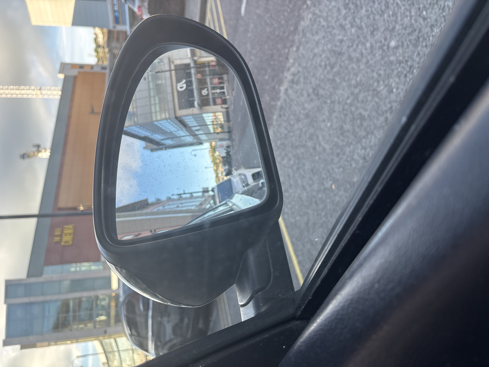 The image is a close-up, low-angle, exterior shot of a car's side mirror, reflecting a street scene. The mirror housing is black plastic and appears slightly dirty. The reflection shows a street with buildings on either side, a white car parked in the middle ground, and a grey car in the background. One building in the reflection has the word "CINEMA" visible in gold lettering. The street surface is asphalt with yellow painted lines. The sky is partly cloudy. The image is taken from inside a car, with the interior of the car visible in the foreground.
Based on the visible signage ("The Reel Cinema" and the architecture), the location is the area around the entrance to The Reel Cinema in Cork, Ireland, which is situated in the city center, likely on a street such as Great George's Street or a nearby road with similar urban development. The specific reflection in the mirror likely captures a street adjacent to the cinema, given the perspective.