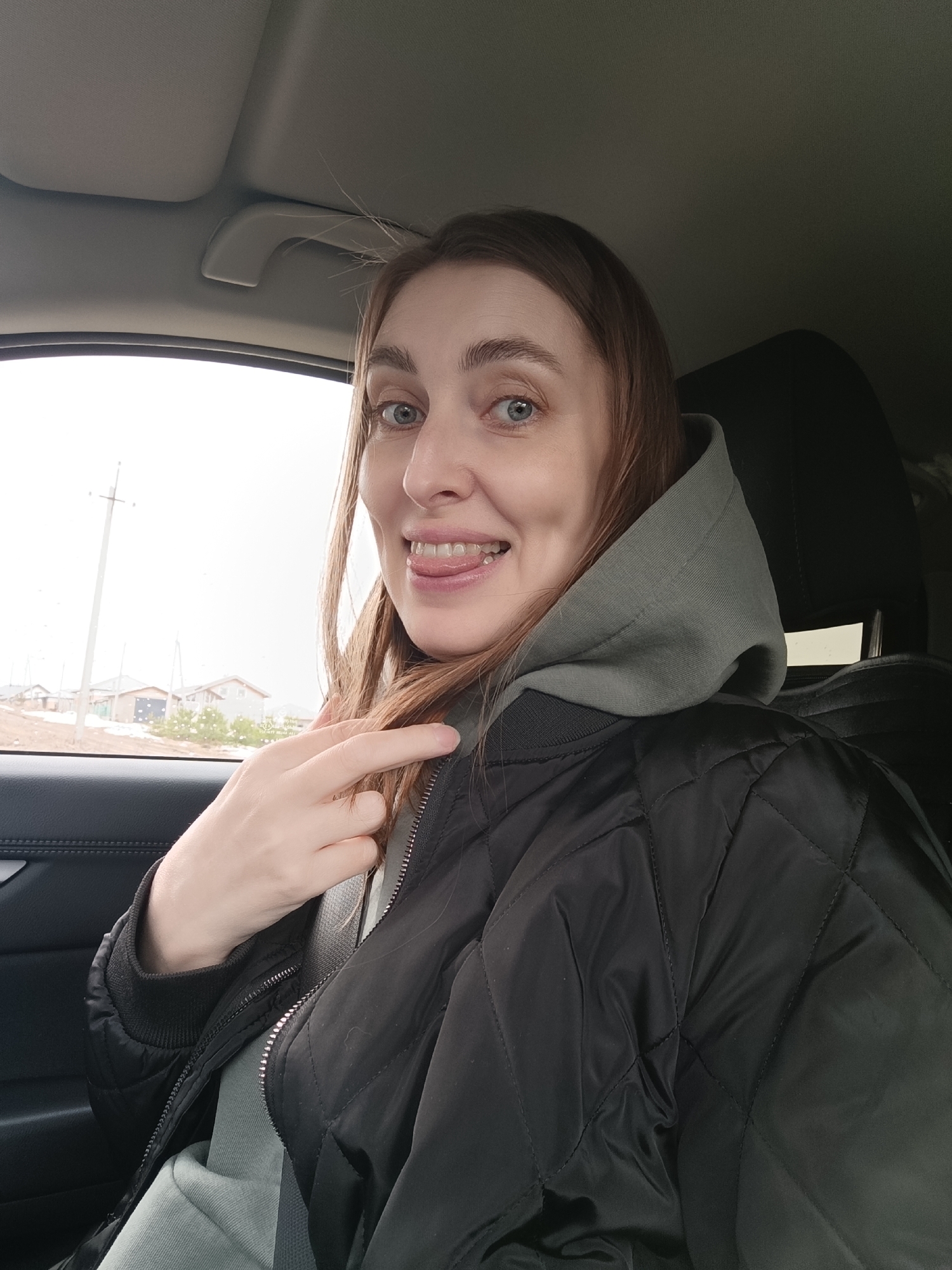 A woman is taking a selfie in a car. She is wearing a black quilted jacket over a grey hooded sweatshirt and is buckled into the seat with a seatbelt. She is sticking her tongue out and pointing with her left hand. Outside the car window, residential houses can be seen with snow on the ground and overcast sky. The location is Chaikovsky, Russia based on the background.