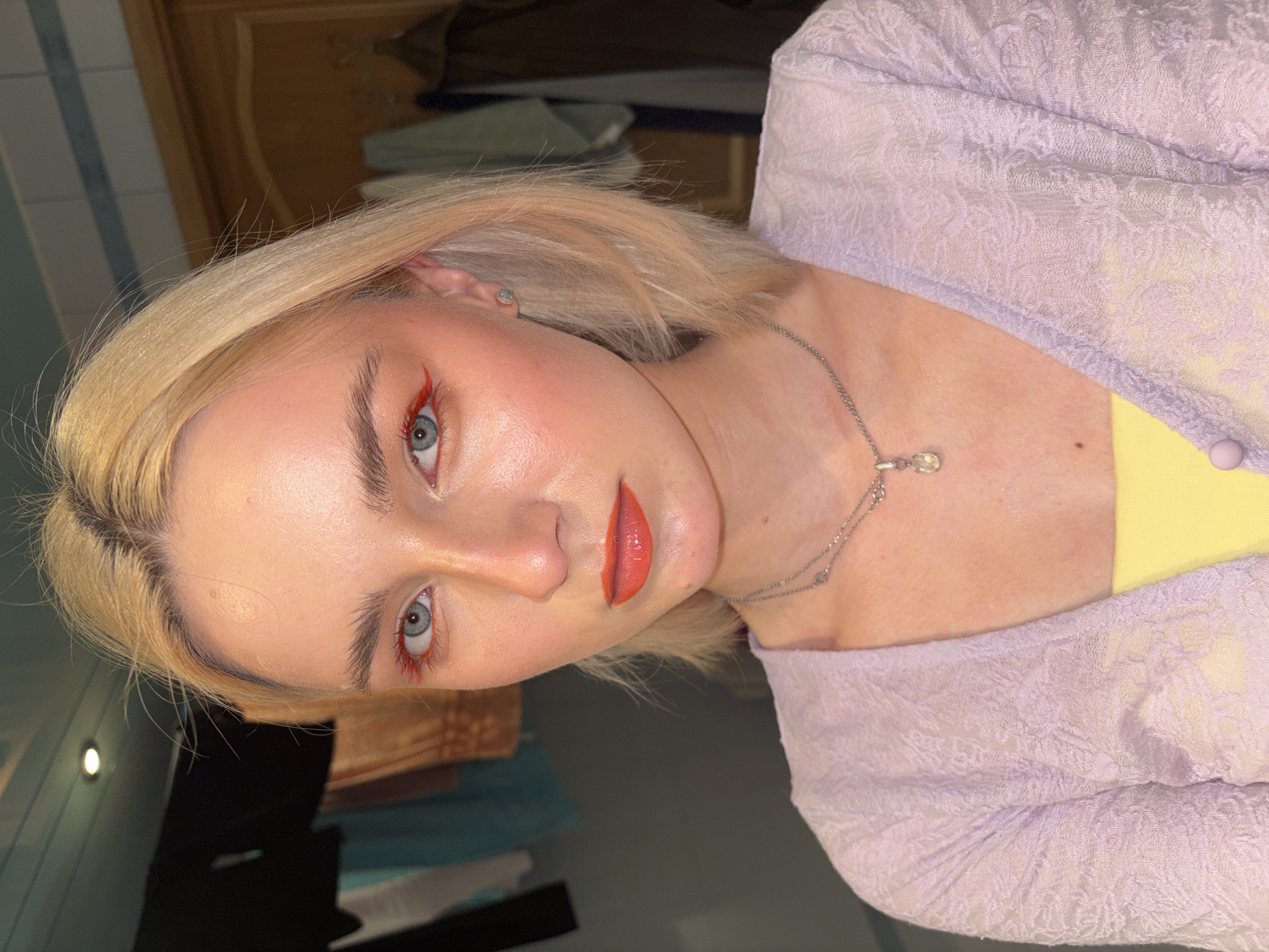 A young woman with blonde, shoulder-length hair and blue eyes is pictured from the chest up, facing forward. She is wearing a pale purple, open-knit cardigan over a yellow top. Her makeup includes vibrant orange-red eyeliner and matching lipstick. She has a delicate silver chain necklace with a clear pendant. The background appears to be an indoor setting with tiled walls and some clothing hanging in the distance. The lighting is bright, creating a slightly reflective sheen on her skin. The overall impression is a close-up portrait with a focus on the woman's makeup and attire. The image does not contain any elements that can be used to determine its location in Sinyavino, Russia.
