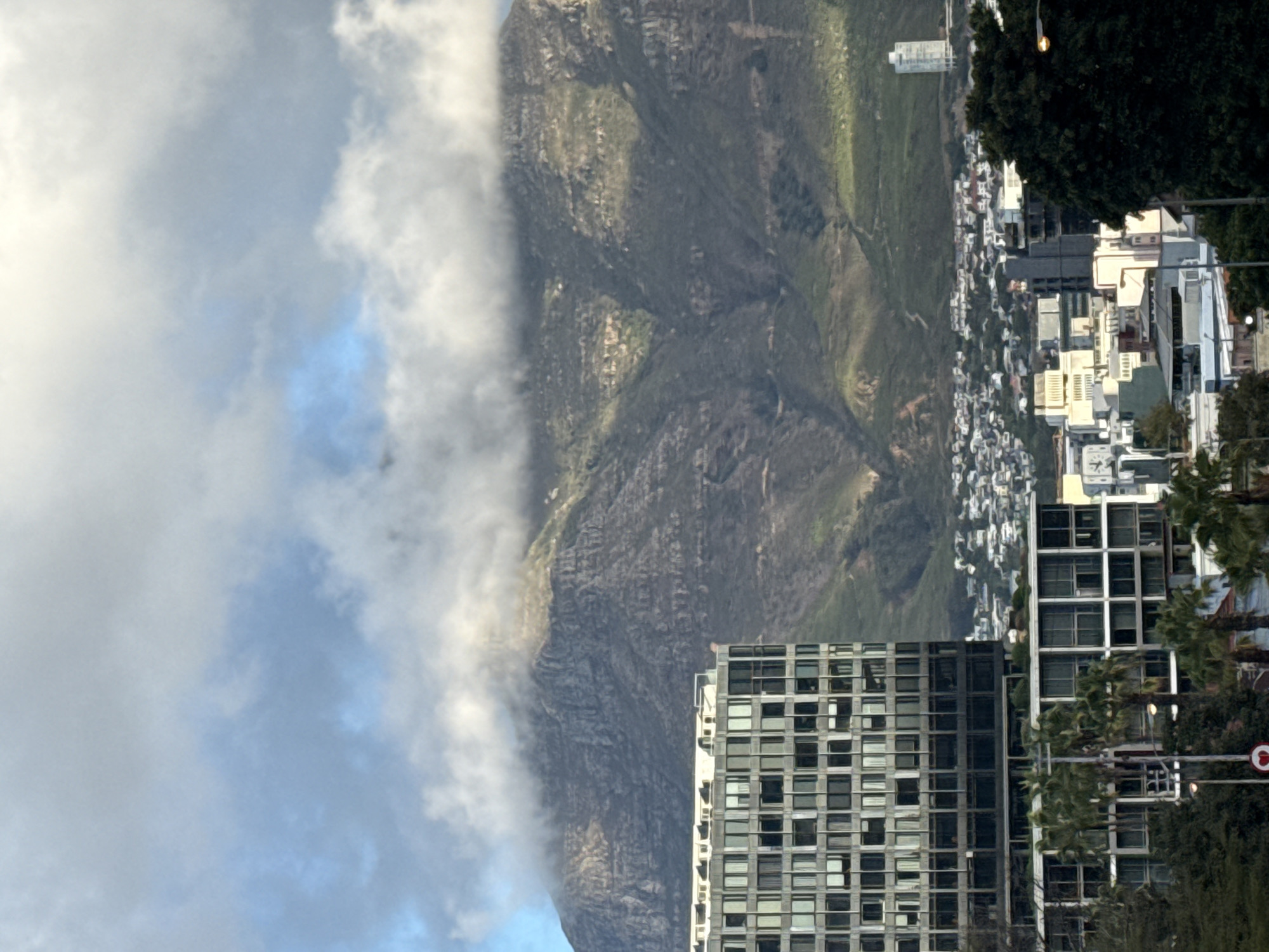 The image depicts a view of Cape Town, South Africa, with Table Mountain in the background partially obscured by clouds. In the foreground is a modern, high-rise building with numerous windows. The building is located near the base of the mountain. Behind the building is a dense urban area with a mix of residential and commercial buildings. The vegetation, including trees and grass, is visible on the slopes of Table Mountain. The sky is overcast with a mix of light and dark clouds.