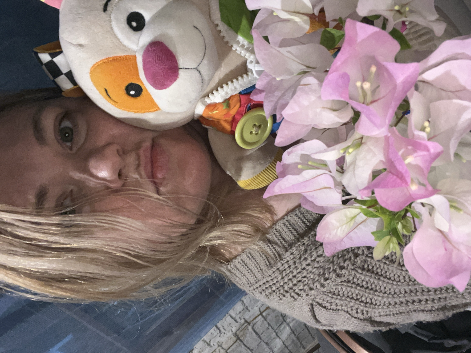 A close-up, vertically oriented image captures a woman alongside a plush toy and a cluster of flowers. The woman, positioned on the left side of the frame, has light-colored hair, part of which falls across her face. Her left eye, light in color, is open and looks towards the viewer. Her nose and mouth, showing slightly parted lips, are also visible. She appears to be wearing a gray knitted sweater, seen on her right arm and shoulder.
To her right, a white and orange plush toy, possibly a cat or similar animal, is visible. It features large black eyes, a pink nose, and an orange snout with a stitched smile. A checkered pattern is visible on an ear flap, and a white zipper with a pull tab reveals colorful fabric underneath, alongside a bright green button.
Dominating the right side of the image is a vibrant cluster of light pink and white flowers, likely bougainvillea, with delicate petals and small, yellowish internal structures. The flowers rest against the woman's shoulder and sweater.
The background is softly blurred, showing hints of a blue surface on the left and a textured, possibly tiled or cobbled, surface below the sweater. The lighting suggests daytime. There is no visible text in the image, and no specific details confirm the location as Vesyoloye, Russia, beyond the context provided. The overall scene appears to be a personal, casual portrait.