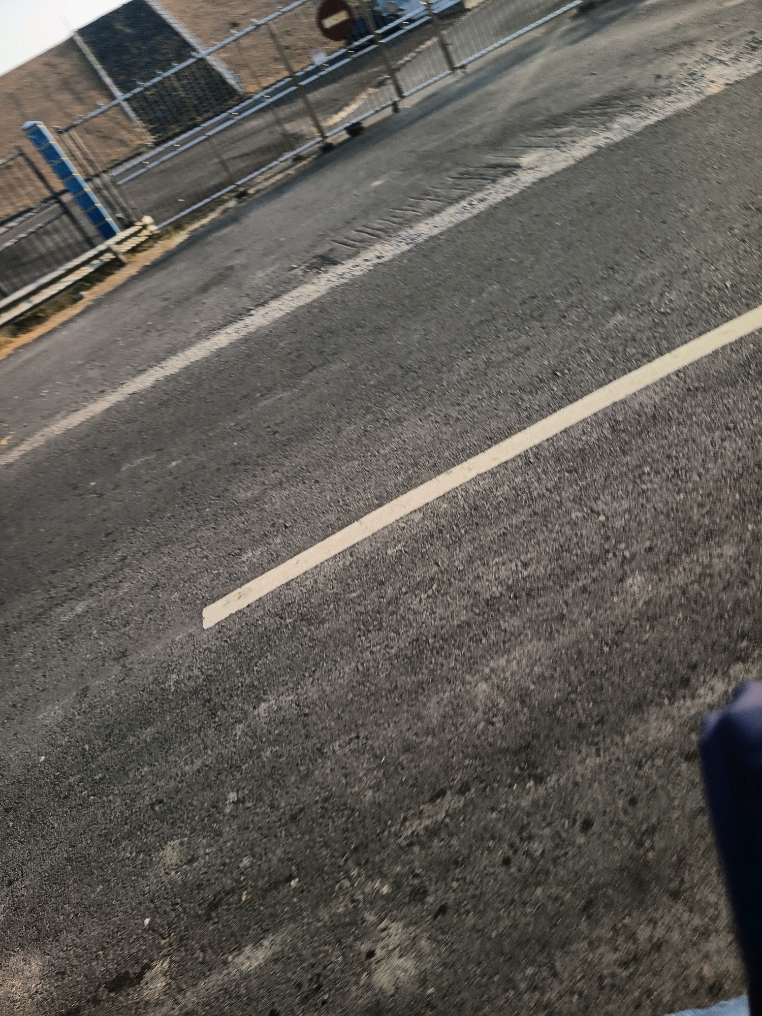 The image shows a close-up, tilted view of an asphalt road with a white line marking its edge. The road surface is textured and appears to be in moderate condition, with some patches of lighter material and subtle depressions. In the background, a metal fence with vertical blue posts is visible, bordering an area with brown, dry vegetation. A circular sign with a red border and a white horizontal bar is mounted on the fence. The overall lighting suggests it is daytime. There are no people or vehicles clearly depicted in the image. The scene appears to be an outdoor environment, possibly a roadside or entrance to an area. No specific details about Kalangbancar, Indonesia, can be identified from the visual information provided.