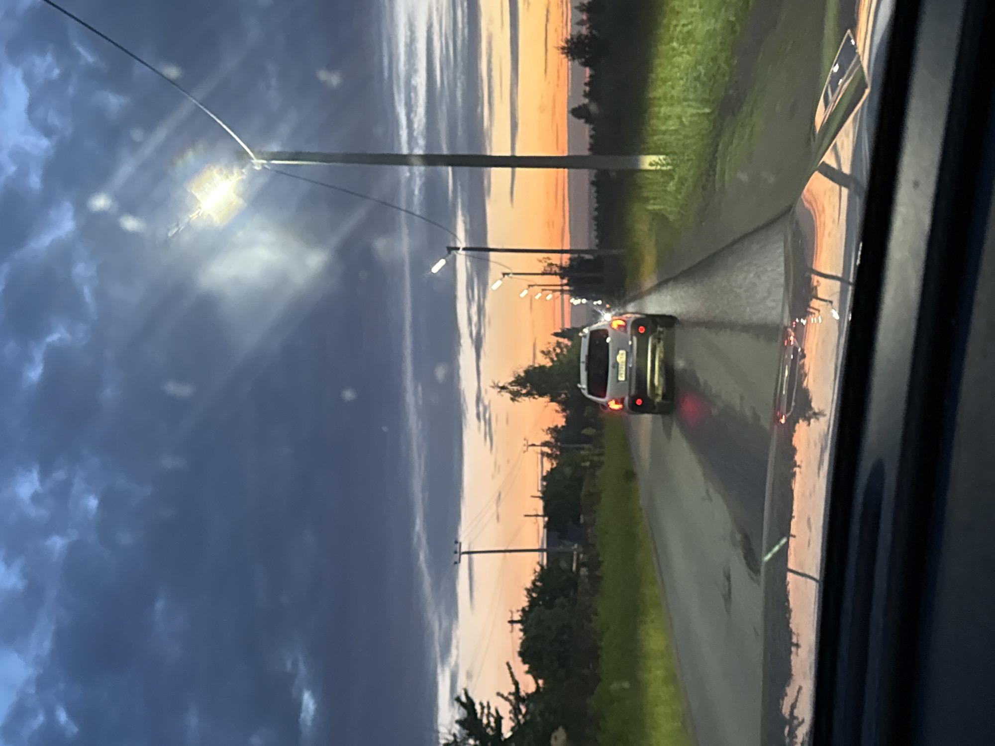 An image taken from inside a vehicle driving on a road during twilight. The road is wet, reflecting the car's lights and the sky. A white SUV is visible ahead, its brake lights illuminated. Streetlights line the right side of the road, casting a bright glow. The sky is a dramatic mix of dark clouds and a vibrant orange and pink sunset. Trees and power lines are visible along the roadside. The dashboard and windshield of the vehicle from which the photo is taken are visible in the foreground, with reflections of the sunset on the windshield. The scene suggests a drive on the outskirts of a city, possibly Kazan, Russia, given the context provided by the prompt.