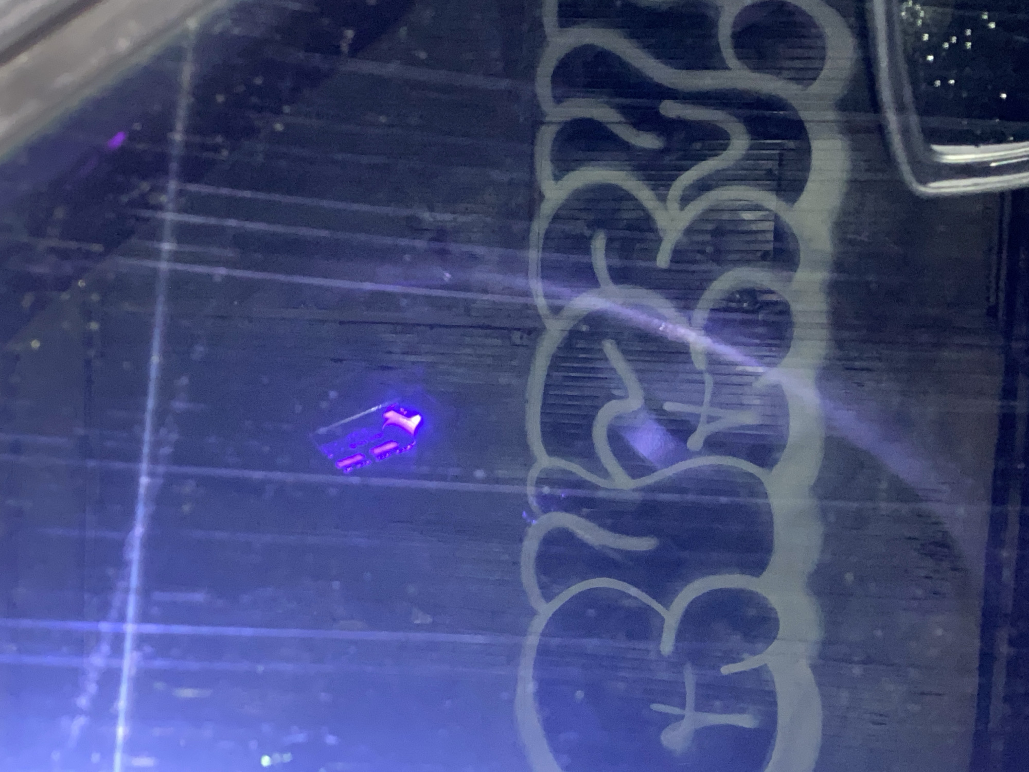 The image presents a close-up, top-down view of a dark, textured surface illuminated by blue or ultraviolet light. The surface features prominent horizontal lines or grooves across its expanse.
A small, rectangular object in the upper-left quadrant emits a bright, intense purple glow. This object appears to be either internally lit or highly fluorescent under the prevailing light, displaying several distinct points of purple illumination within its form.
Extending vertically along the right side of the image is a light-colored, stylized pattern resembling graffiti or street art. This design consists of thick, curvy, abstract lines and shapes; while artistic, no specific readable text or words are clearly discernible.
Numerous tiny, bright specks, possibly dust, reflections, or debris, are scattered across the entire blue surface. The lighting conditions are low, characterized by a dominant blue and purple hue, suggesting the use of artificial light, potentially a UV lamp, which causes the object to fluoresce. Faint light streaks and reflections are also visible, particularly towards the bottom and center of the frame.
No people or active events are depicted; the image captures a static arrangement of objects and patterns within what appears to be an indoor setting, possibly an art display or a casually lit environment.