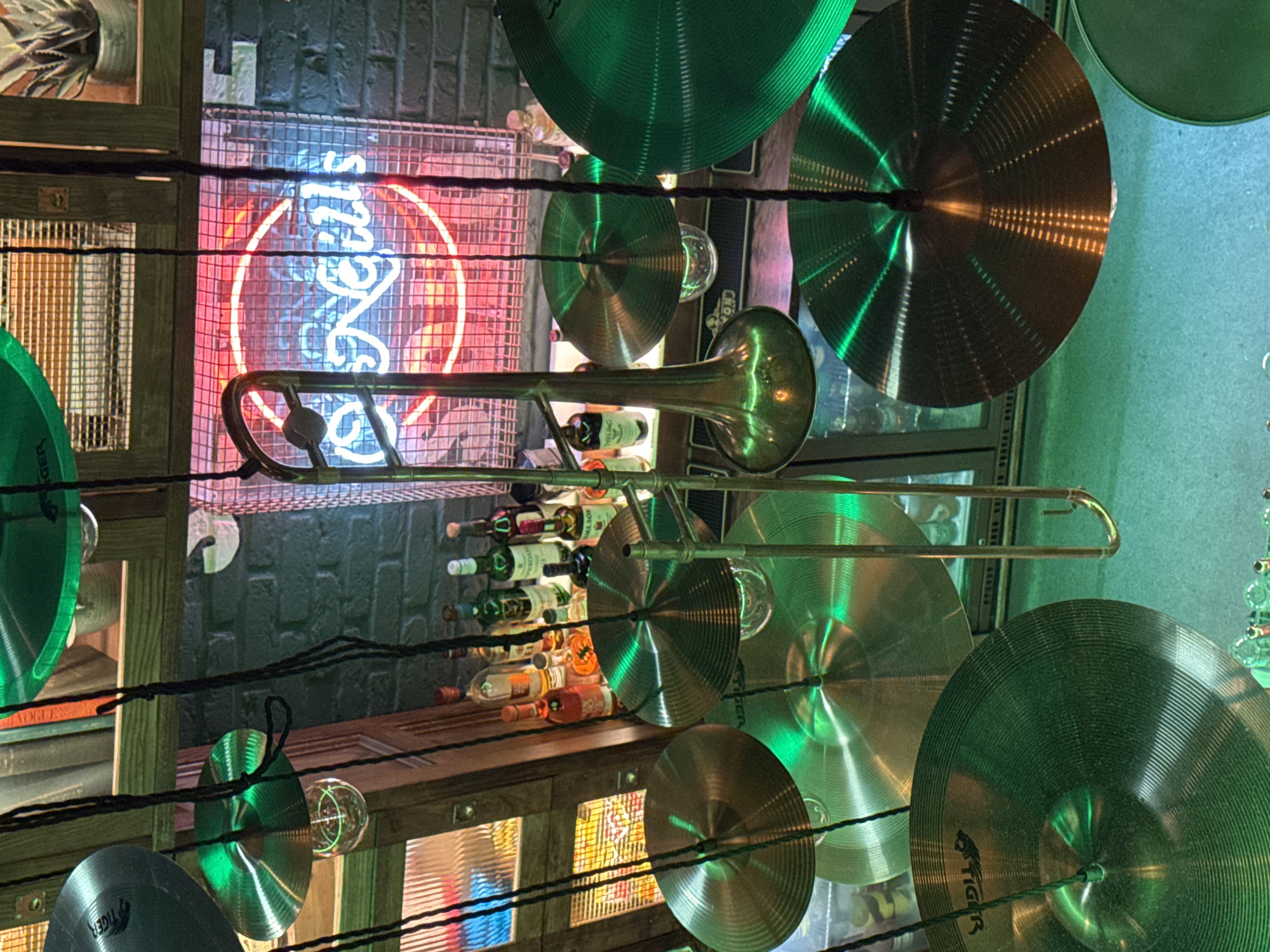 The image displays a creatively decorated indoor space, likely a bar, restaurant, or music venue with a distinct jazz theme. Numerous cymbals, predominantly with a green tint from ambient lighting, are densely suspended from above, forming a unique ceiling or decorative screen. Several of these cymbals feature the brand name "TIGER".
A golden trombone is prominently positioned horizontally across the mid-ground, nestled amongst the hanging cymbals. Its reflective surface gleams under the soft lighting. In the background, a striking neon sign glows with red and blue lights, forming a circular outline and lettering that appears to spell "OYSTERS". This sign is set against a dark brick wall.
Below the neon sign, a bar area is visible, featuring shelves stocked with various bottles of spirits and wine. Labels such as "JAMESON" and "TEELING" can be faintly identified on some of the bottles. Additional decorative elements include exposed light bulbs interspersed with the cymbals and wooden structural components. The overall lighting suggests an intimate, evening or nighttime atmosphere. No people are visible in the scene. The decor strongly implies a place for live music and drinks.