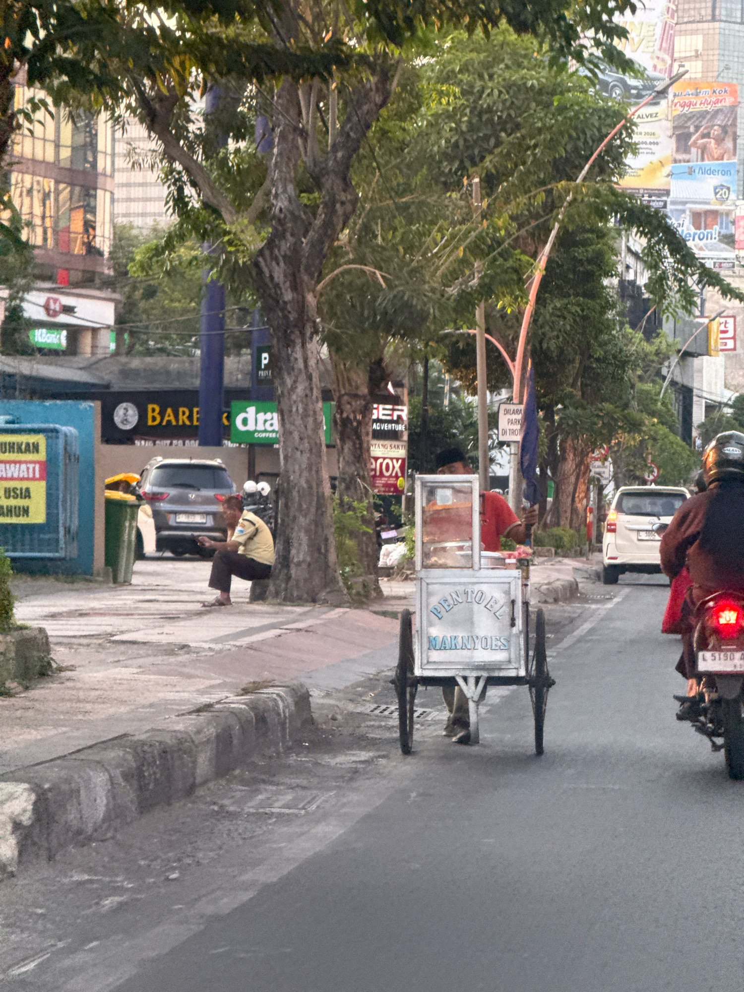 The image depicts a street scene in Surabaya, Indonesia, with a focus on a food cart vendor. In the foreground, a white food cart with the words "PENTOEL MAKNYOES" written on it is positioned on the road. The cart has two wheels at the back and a single wheel or support at the front. A person wearing a red shirt and dark pants is standing behind the cart, appearing to be the vendor.
To the right of the cart, a person is riding a motorcycle, seen from the back. The motorcycle has a license plate that reads "L 5190". The rider is wearing a helmet and a dark jacket, with a glimpse of a red garment underneath.
On the left side of the street, a man in a yellow and brown uniform, possibly a security guard or traffic officer, is sitting on the curb next to a green trash bin. He is looking down at his phone. Behind him, a gray car is parked.
The background features trees lining the sidewalk, with tall buildings and billboards visible further in the distance. The lighting suggests it is either early morning or late afternoon. The environment appears to be a moderately busy urban street.