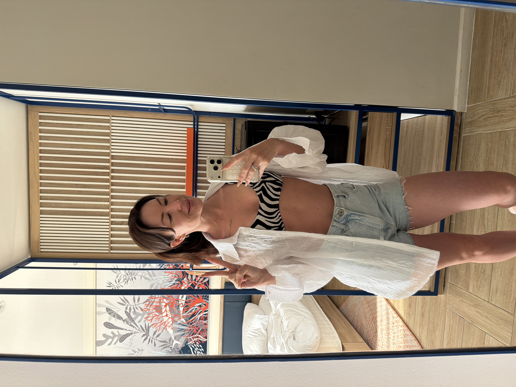 A woman is taking a mirror selfie in a room. She is wearing a zebra-print bikini top, light-wash denim shorts, and an unbuttoned white linen shirt. She is holding her phone in her right hand and making a peace sign with her left hand. In the background, a bed with white bedding is visible, along with a wall decorated with a coral and seaweed mural. The room has light wood flooring and a blue-framed shelving unit. The overall scene suggests a relaxed or vacation setting. There is no indication that the location is the city of Dong Ninh Hoa Ward, Vietnam.