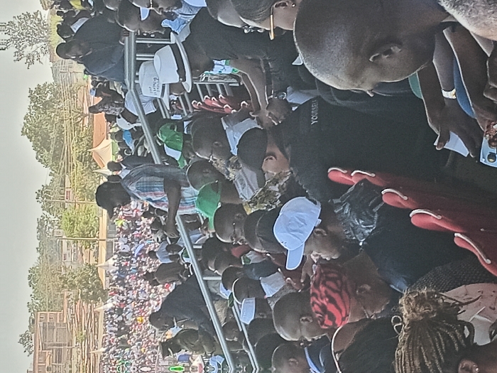This media file captures a large outdoor gathering of people, likely a public event such as a rally or meeting, set in Siaya, Kenya. Hundreds of individuals, predominantly adults and appearing mostly male, fill the frame, extending into the background.
In the foreground and mid-ground, people are standing and sitting on various structures, including what appear to be temporary metal barriers or bleachers. Many are casually dressed in shirts and t-shirts. Some individuals wear hats; one notable detail is a white fedora-style hat on which the text "FREEDOM OF SPEECH" is clearly visible. Another person in a black t-shirt has partially visible text that appears to read "YOURSELF". Red plastic chairs are visible in some sections, some occupied, others holding personal items.
The crowd is oriented towards a central, unseen point, suggesting a speaker or performance. In the background, beyond the immediate gathering, rows of people are visible, indicating the vastness of the crowd. Further back, there are trees, simple buildings, and a large white tent, possibly for shade or as a designated area. The overall lighting suggests it is daytime, likely late morning or afternoon, with clear weather conditions. The atmosphere appears attentive and engaged.