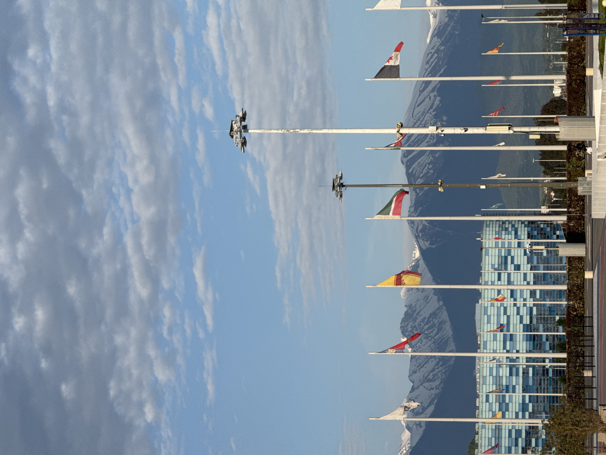 The image shows a row of flagpoles with various flags waving in the wind, set against a backdrop of mountains with visible snow-capped peaks. The sky is partly cloudy. The flags are positioned in front of a modern, multi-story building with a blue and white grid-like facade. The location appears to be in the city of Sirius, Russia, indicated by the architectural style of the buildings and the mountainous background, possibly near the Black Sea coast. The image is oriented sideways.