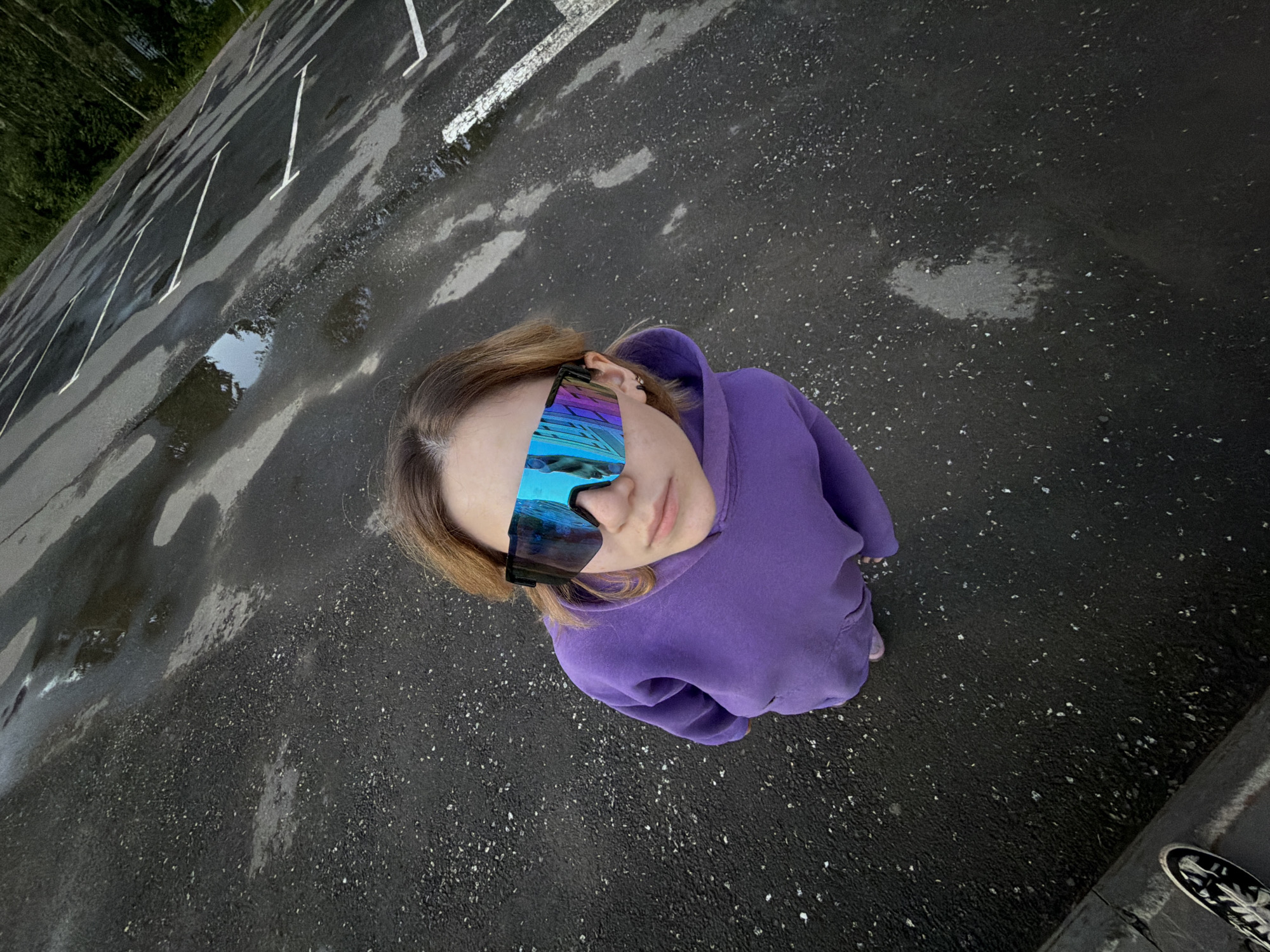 The media file depicts a young person standing in an outdoor parking lot. The subject is viewed from a high angle, looking directly up at the camera. They are wearing a vibrant purple hoodie and striking, reflective, wrap-around sunglasses that mirror a blue sky with white clouds. The parking lot surface is dark asphalt, wet and reflecting the surrounding environment, with several white parking lines visible. Puddles of water are scattered across the lot, indicating recent rain. In the background, to the left, a dense green forest can be seen. The overall impression is of a casual, possibly post-rain moment. The lighting suggests it could be daytime, given the bright reflections in the sunglasses, though the sky in the reflections is cloudy. There are no visible text elements in the image.