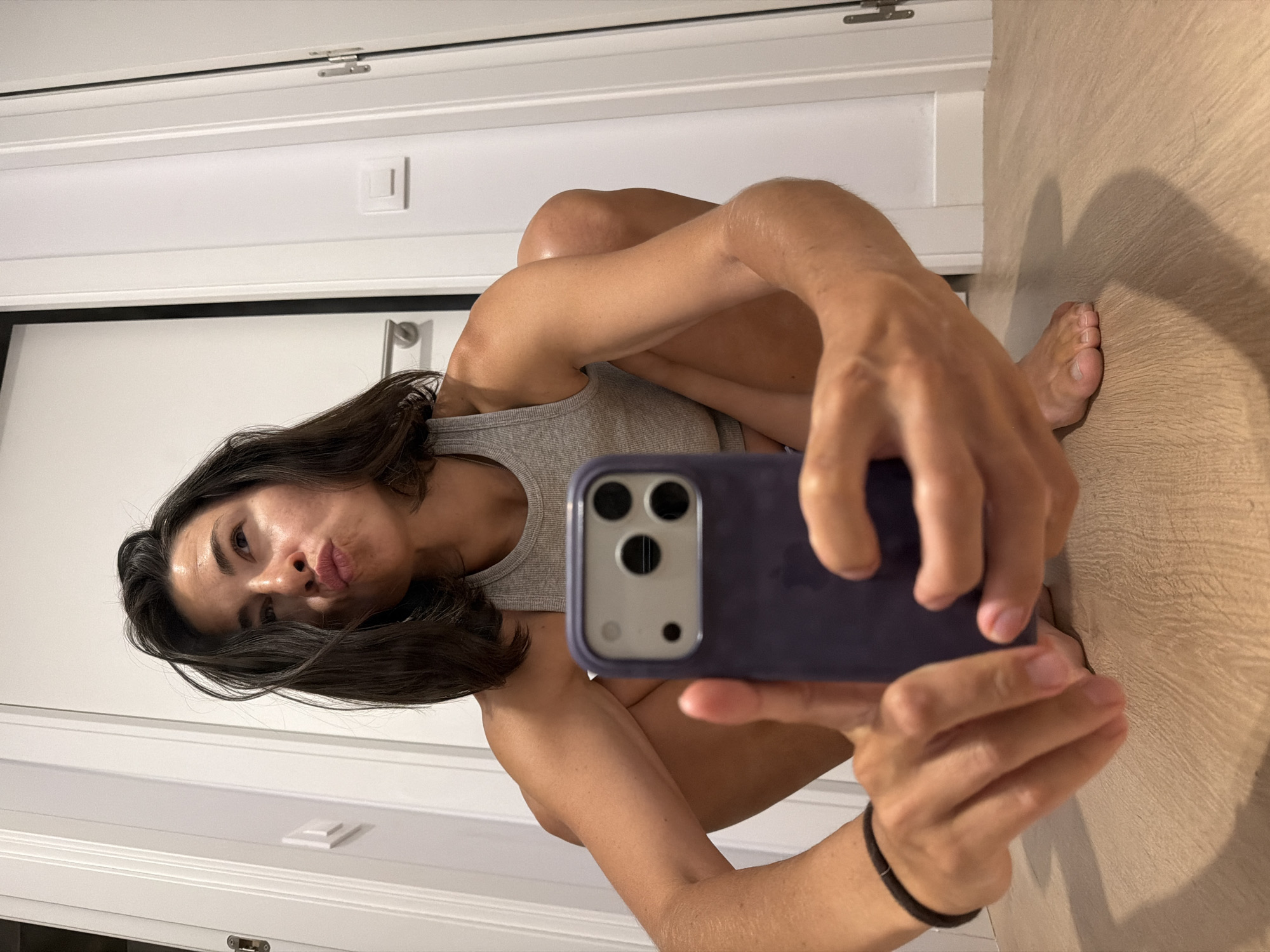 A young adult woman is in a low crouch indoors, facing the camera and taking a selfie with her phone. She has dark hair, pulled back from her face, and is wearing a grey tank top. Her left arm is bent, holding the phone, and her right arm rests on her right leg. The background appears to be a hallway with white doors and a light-colored wooden floor. The setting is consistent with an apartment or residential building in l'Hospitalet de Llobregat, Spain, characterized by modern interior design and common architectural elements found in urban residential areas. The lighting is natural, suggesting daylight coming from an unseen source.