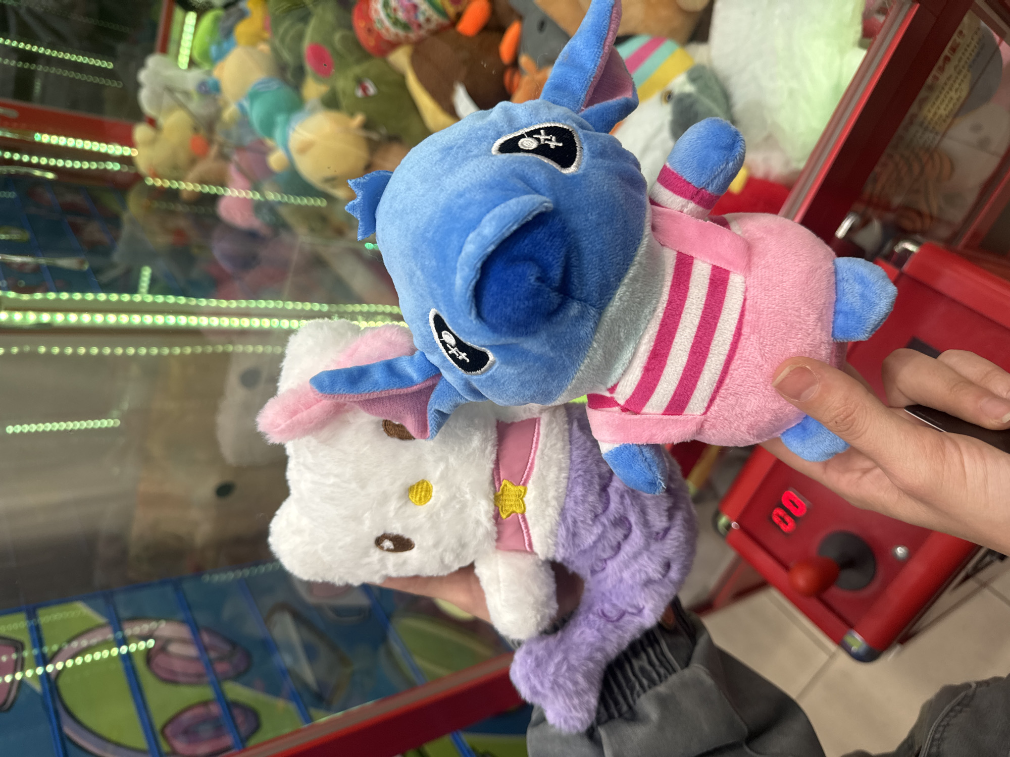 In an indoor setting, likely an arcade or entertainment center in Krasnoyarsk, Russia, a person's hands are holding two plush toys. The top plush resembles Disney's Stitch, characterized by its blue color, large ears with pink inner lining, and embroidered eyes with white cross-stitches on black. It wears a pink outfit with white and fuchsia stripes.
Below the Stitch plush is a white plush toy, reminiscent of Hello Kitty, featuring embroidered brown eyes and nose, and a small yellow star on a pink harness around its body. Uniquely, this plush has a textured purple mermaid tail.
In the background, behind the held toys, is the interior of a claw machine. Various other plush toys, including yellow, green, and brown ones, are visible inside. Green string lights illuminate the edges of the machine's glass casing. To the right of the person's hands, part of a red arcade game console is visible, equipped with a red joystick and a digital display showing "00", likely indicating remaining credits or a score. The overall impression is that the plush toys have just been won or are being held near the game.