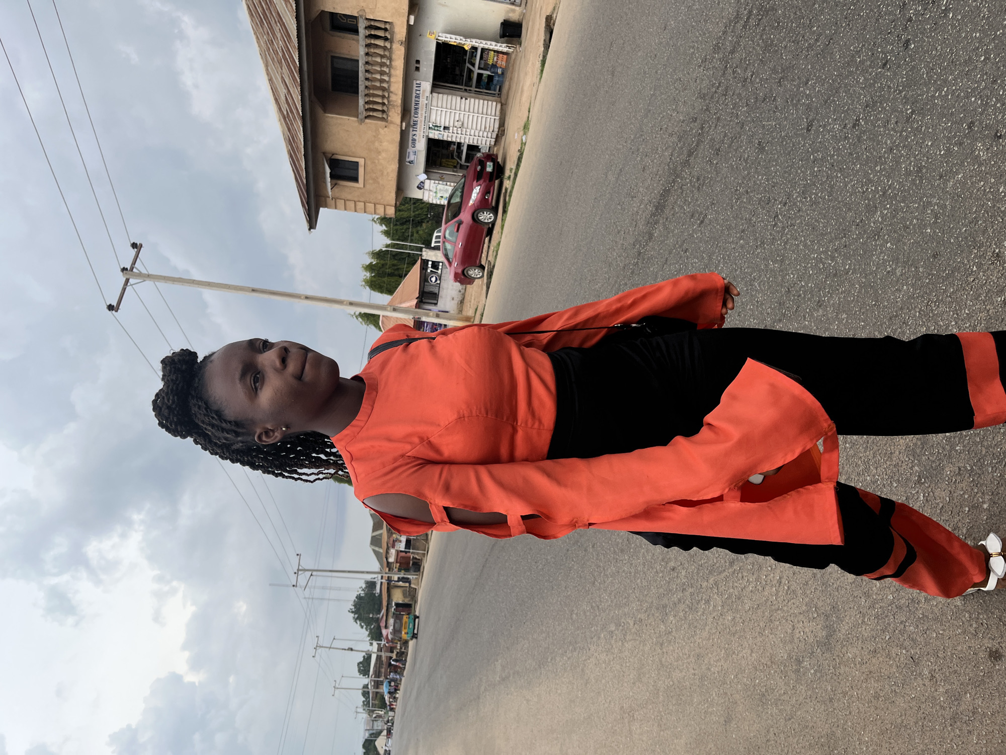 A young Black woman with braided hair styled in a bun walks along the side of a paved road in Barakin Lerawi, Nigeria. She is wearing a vibrant orange, long-sleeved shirt with cutouts on the sleeves, and black pants with orange accents. She has on white sandals with a gold buckle. The road is lined with buildings, some appearing to be shops, and utility poles with electrical wires. A maroon sedan is parked on the side of the road in front of a building labeled "GOD'S TIME COMMERCIAL". In the background, a yellow auto-rickshaw is visible further down the road. The sky is overcast.