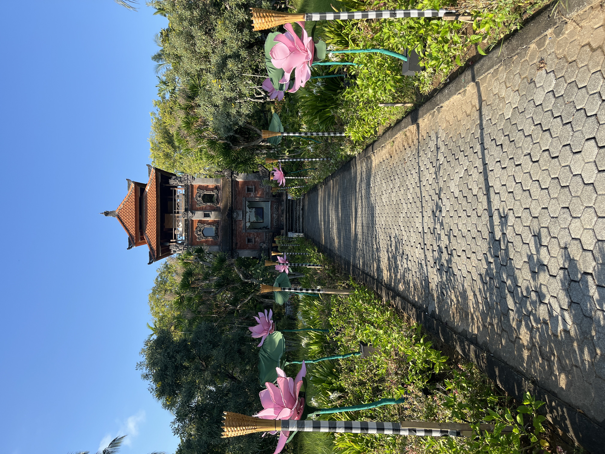 An exterior, eye-level, full shot shows a pathway leading to a traditional Indonesian building. The pathway is paved with hexagonal gray stones and lined with lush green bushes and decorative elements resembling large pink lotus flowers. Each lotus is mounted on a green pole with a woven torch-like fixture at the top, and many are adorned with black and white striped bands. The building in the background, possibly a temple or traditional house, features a tiered, red-tiled roof and intricate stone carvings. It is surrounded by abundant green trees and foliage under a clear blue sky. The scene is bathed in daylight, casting shadows from the decorative elements and trees onto the pathway.