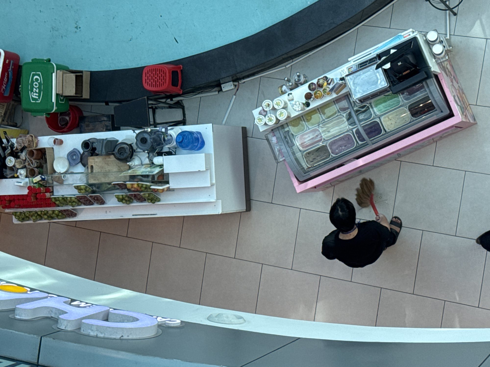 An overhead shot captures an outdoor food stall, likely located in Da Lat, Vietnam, characterized by its cool climate and abundance of flowers. The stall features a pink-trimmed ice cream display case filled with various colorful flavors. To the right of the ice cream case, a white shelf holds several small cups with toppings. In the background, a longer white counter displays an assortment of fruits in trays, alongside containers, a blender, and a blue water jug. A person in a black shirt and pants is actively sweeping the tiled floor with a red-bristled broom. Adjacent to the longer counter, a green cooler labeled "Cozy" is visible, as is a small red stool. The scene unfolds on a light-tiled floor with dark grout lines, adjacent to a blue body of water.