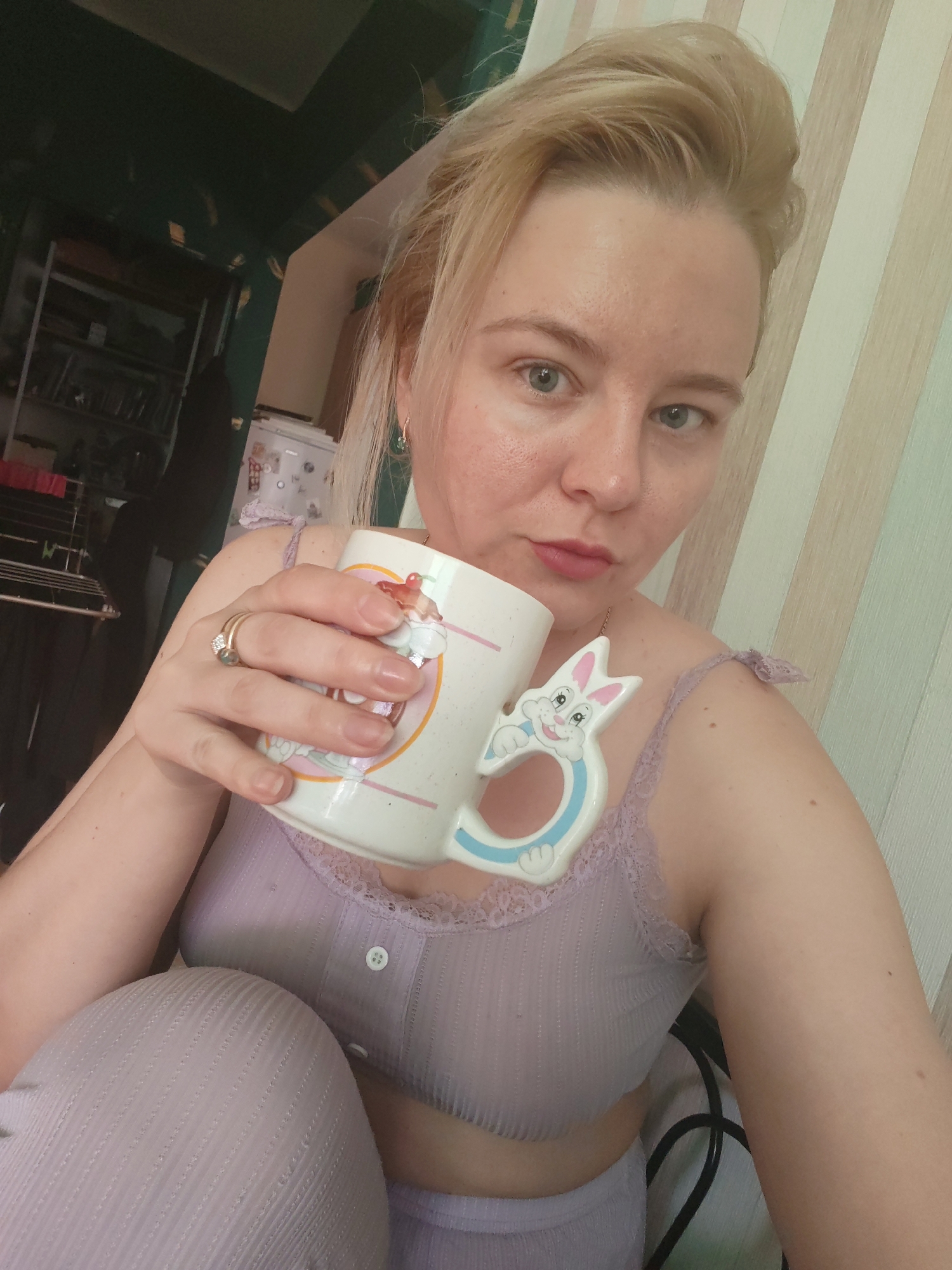 A medium eye-level shot depicts a woman with fair skin, blond hair pulled back, and gray eyes, holding a white ceramic mug with a cartoon bunny handle. She wears a light purple top with lace trim and a button-up front, paired with matching bottoms. The background includes striped wallpaper and a glimpse of a room with shelves and various items. The location is Izhevsk, Russia.