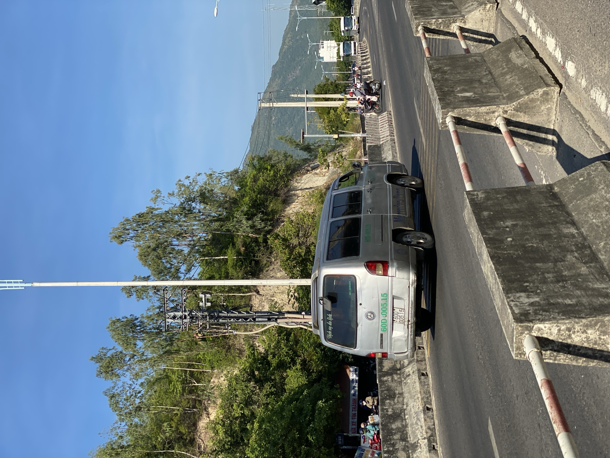 In an outdoor, daytime scene, a silver van with a license plate reading "60D.005.15" drives on a multi-lane road, separated by concrete barriers. The van has a Mercedes-Benz logo on the rear. The road is flanked by a steep, tree-covered embankment on the left and a mountainous landscape under a clear blue sky on the right. Other vehicles, including motorcycles and buses, are visible in the distance on the road. Power lines and utility poles are present. Several people are seen on motorcycles near the embankment.