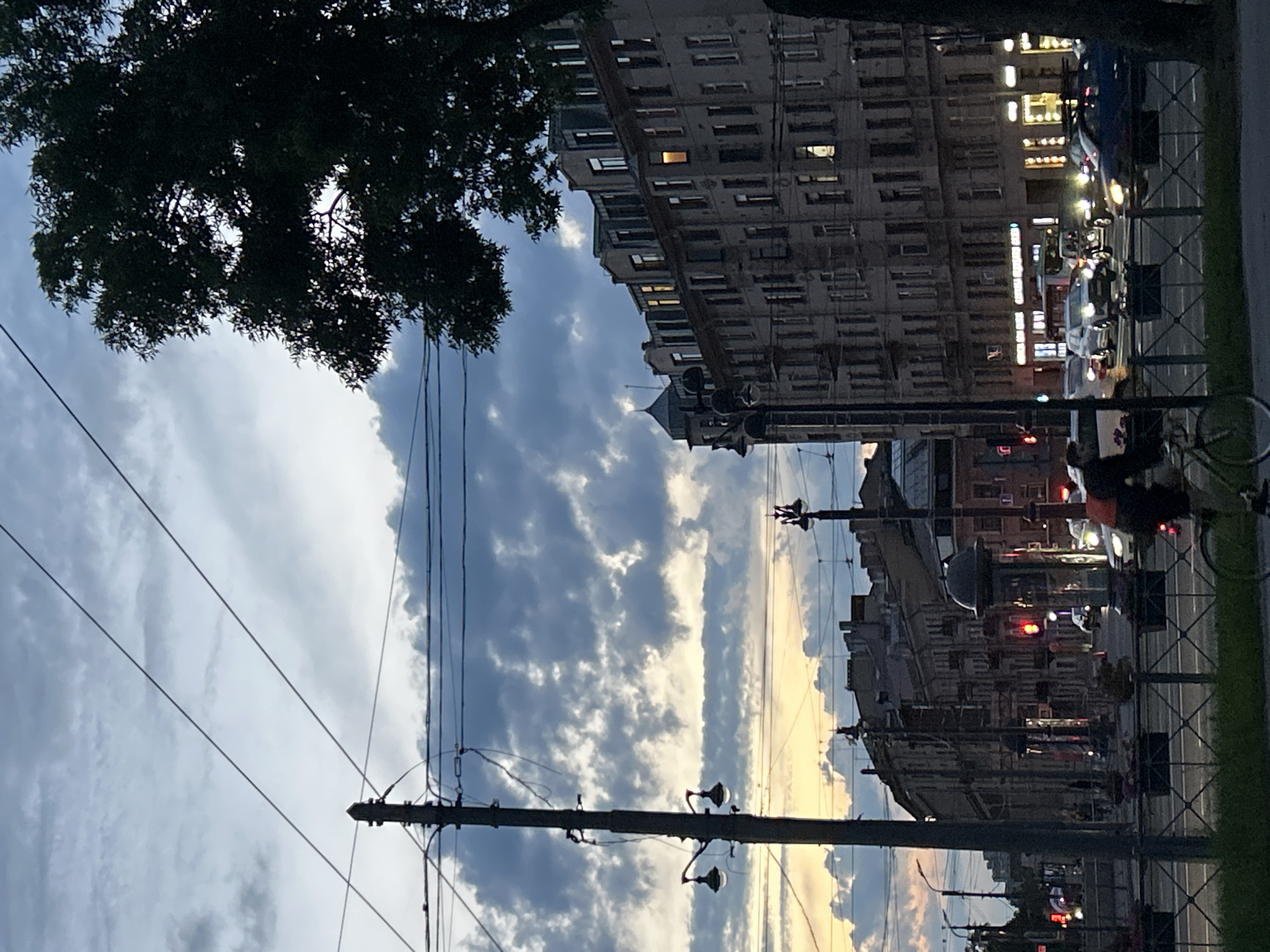 In an urban street scene at dusk, a four-story building with mansard roofs and numerous windows lines the right side of the frame. Some windows are illuminated, suggesting interior lights. A large tree with dark foliage is silhouetted against the sky on the far right. Overhead, utility wires crisscross the scene. A tall utility pole with a street lamp stands to the left of center, with another street lamp visible further down the street. In the foreground, a person on a bicycle is moving away from the viewer. Several cars are on the street, some with their headlights on, indicating traffic. The sky is filled with dynamic clouds, with a band of orange and yellow light visible near the horizon, suggesting the last rays of sunset. The buildings in the background appear to be of similar architectural style to the main building. The scene is captured at a slight downward angle, looking towards the street and buildings.