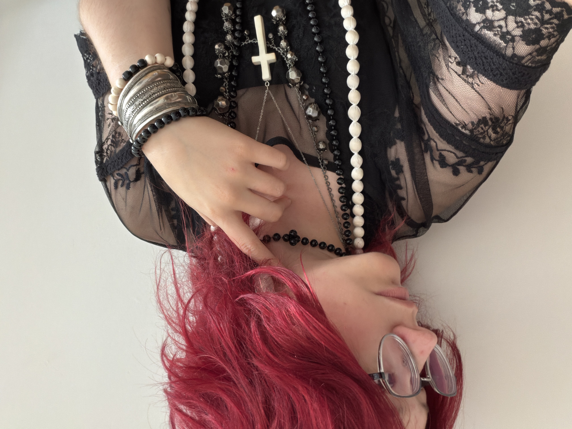 The image shows a close-up of a person with vibrant red hair and glasses. The person is wearing a black, sheer lace top with floral patterns. They are adorned with multiple necklaces, including a black choker with a floral design, a longer silver chain with a white cross pendant, and strands of black and white beads. On their left wrist, they are wearing several bracelets, including a wide silver cuff bracelet layered with black and white beaded bracelets. The person is looking to the left, with their right hand gently touching their neck. The background is a plain, light-colored wall. There is no information in the image to suggest a location in Chelyabinsk, Russia.