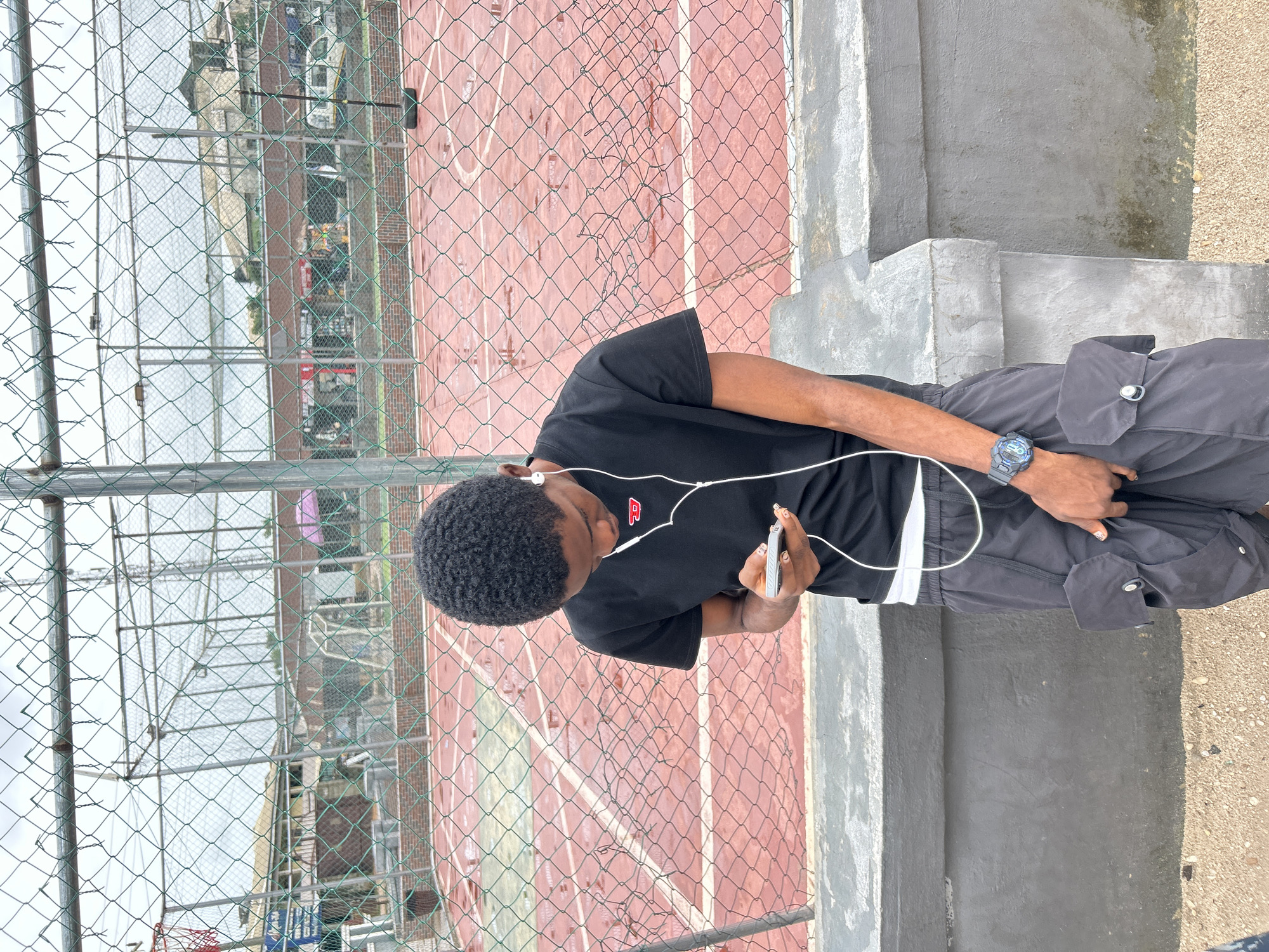 A young Black male, wearing a black t-shirt, grey cargo pants, and a blue digital watch, stands in front of a chain-link fence. He is looking down at his phone, which he holds in his left hand, and has white wired earphones in his ears. The fence separates him from a red basketball court with white lines. Beyond the court, there are buildings and trees, characteristic of the city of Ihiagwa, Nigeria. The ground in front of the fence is covered in gravel. The lighting suggests it is daytime, possibly overcast.