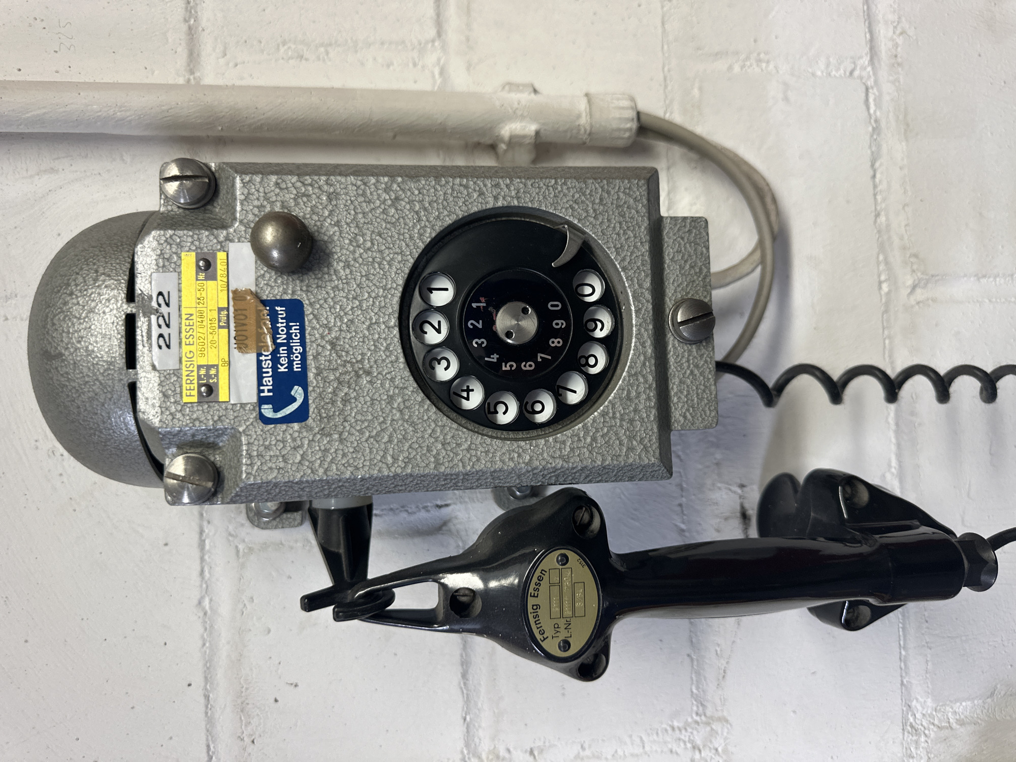 This is a medium shot, eye-level, outdoor photograph of an old, rotary dial telephone mounted on a white brick wall. The telephone is grey metal, with a black handset hanging below. The telephone has a round dial with black numbers on a white background, and a metal rotary finger stop. A coiled black cord connects the handset to the main unit.
Visible text on the telephone includes labels with "FERNSIG ESSEN" and "Haustel Kein Notruf möglich!" which translates to "No emergency call possible!". There are also serial numbers and model information visible on small labels.
The telephone appears to be an older model, perhaps from the mid to late 20th century, suggesting a historical or utilitarian setting. The white brick wall and a white pipe running above the telephone suggest an industrial or institutional environment. The overall impression is one of vintage technology and functionality, possibly in a location where standard telephone service was limited or unavailable for emergency calls. The lighting suggests it's daytime. There are no people visible in the photograph.