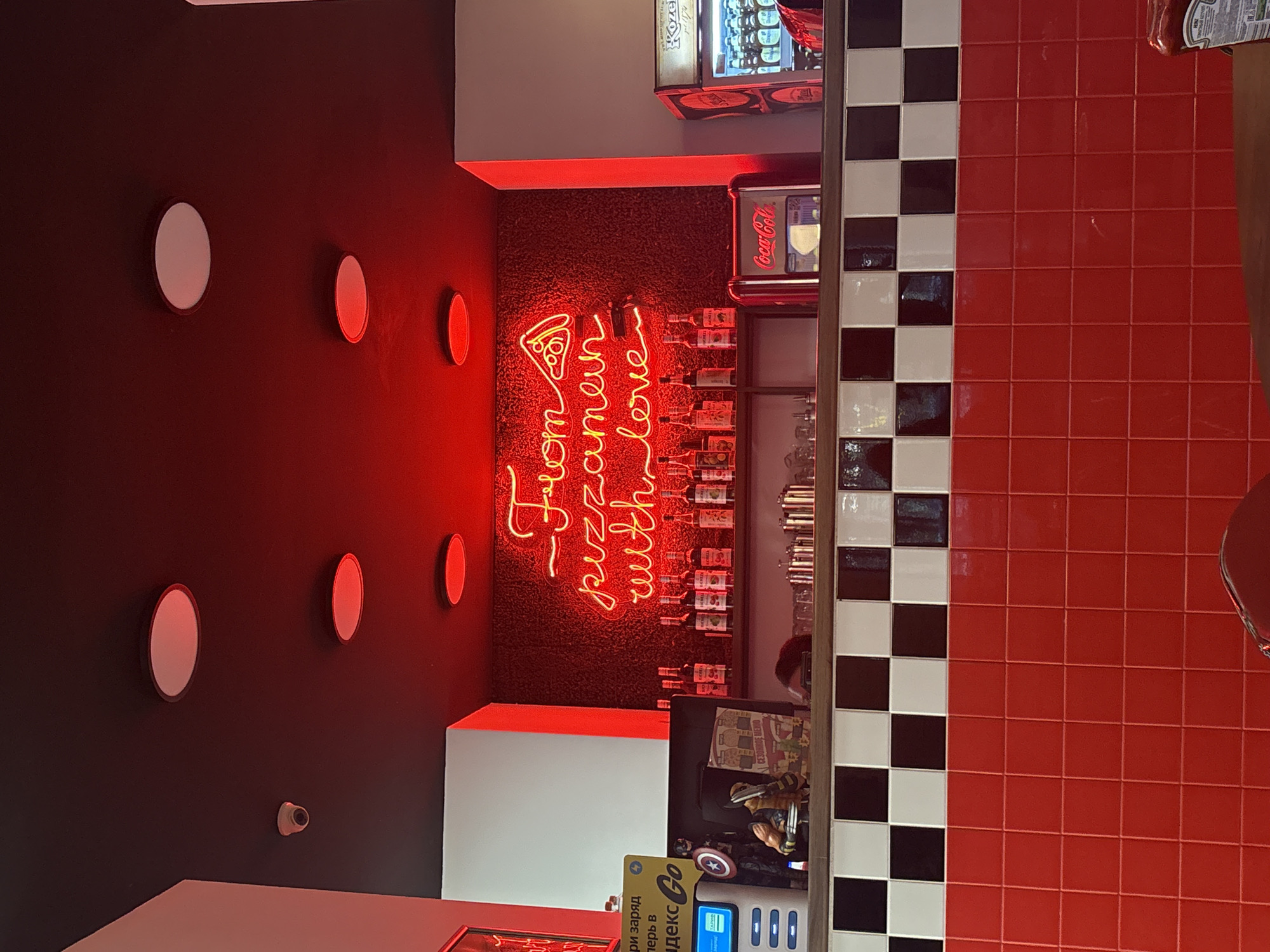 The image shows the interior of a pizza establishment, likely located in Perm, Russia based on the signage and branding. A black wall is adorned with several round, illuminated white and red fixtures. In the center is a neon sign that reads "From pizzamen with love," accompanied by a pizza slice graphic. The area above the sign features shelves stocked with bottles, likely ingredients or condiments. Adjacent to this section is a wall tiled with alternating black and white squares and a larger area of red square tiles. A Coca-Cola branded vending machine is visible, along with another vending machine with the "Koze 1874" brand, indicating beverages for sale. On the lower right, a metallic table leg is visible. A stand or small kiosk is visible at the bottom of the image, featuring "Яндекс Go" branding, suggesting a point for online orders or service. Overall, the scene conveys a retro diner or pizza parlor vibe with a modern, branded twist.