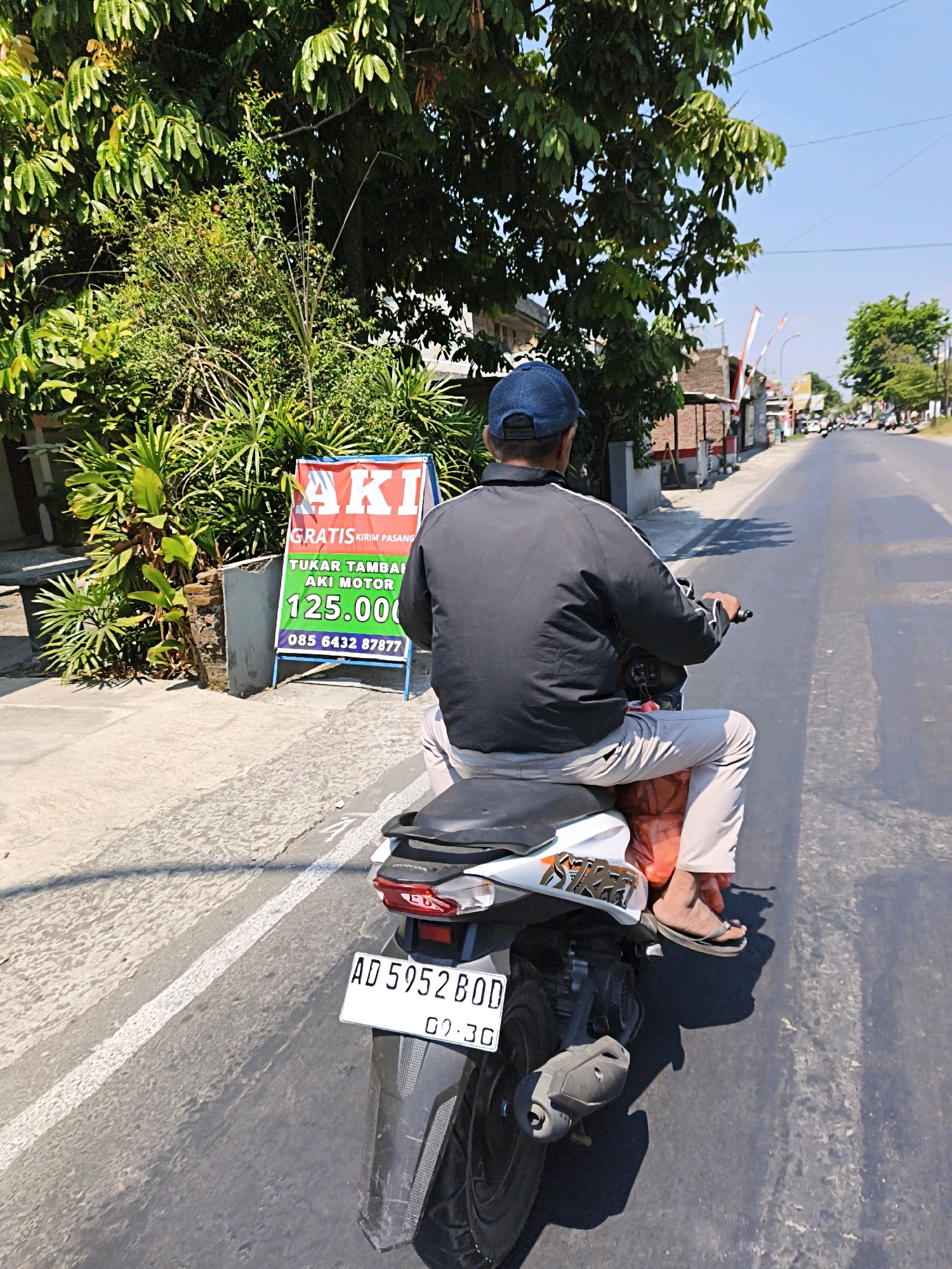 A man wearing a blue baseball cap and a black jacket rides a white and black motorcycle on a paved road in Desa Donohudan, Indonesia. The man is wearing light-colored pants and dark flip-flops. He is carrying a large orange plastic bag on the back of his motorcycle. To the left of the road is a sign in red, white, and green that advertises "AKI MOTOR" (motorcycle battery) with a price of "125.000". Behind the sign are lush green trees and plants. In the background, there are buildings, other motorcycles, and trees. The sky is clear and blue. The image is taken from behind the rider, looking forward.