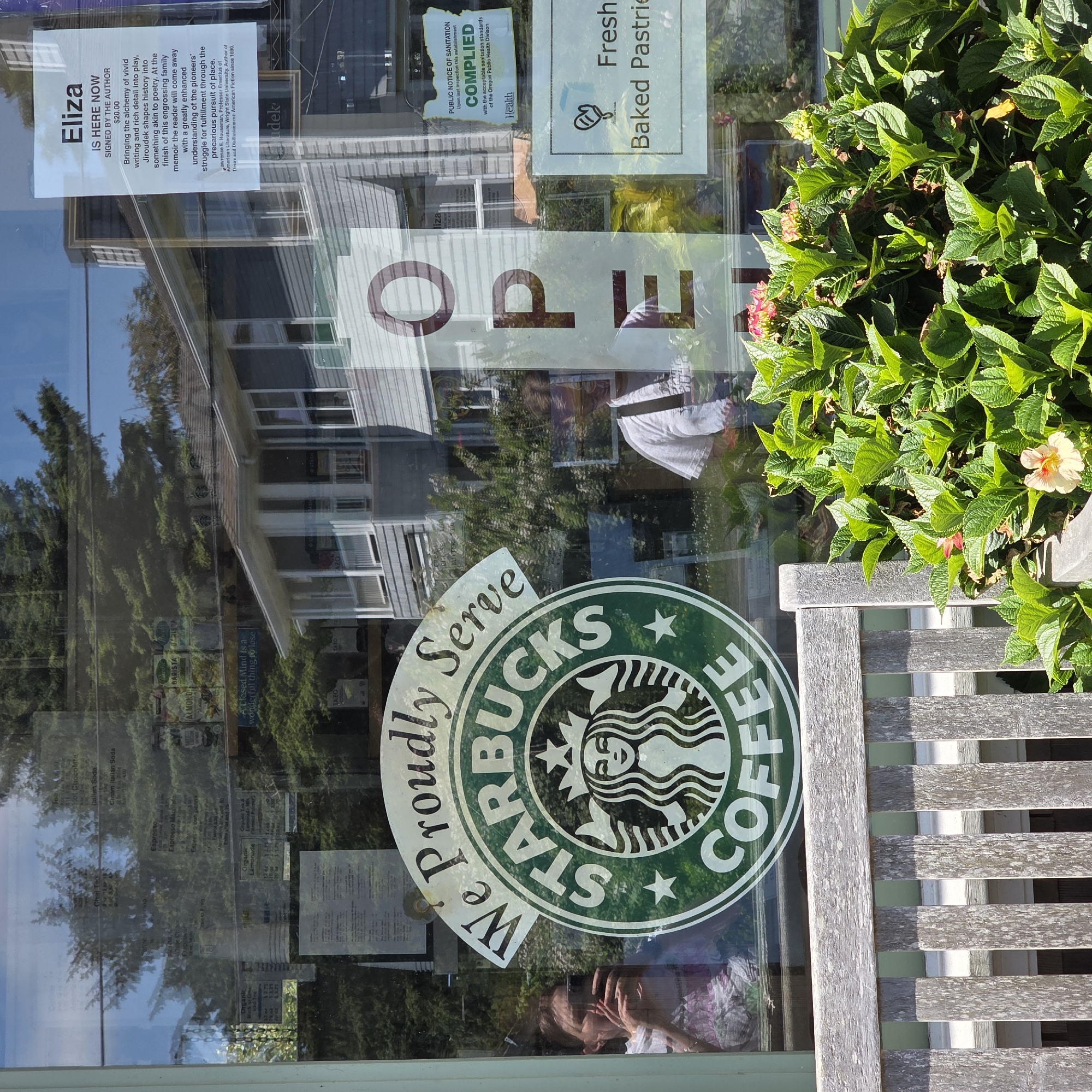 The image depicts a street-level view of a Starbucks store in Cannon Beach, Oregon, United States of America. The store's exterior window displays a large Starbucks logo with the text "We Proudly Serve Starbucks Coffee." The window reflects trees, buildings, and sky. A woman is visible through the window, taking a photo with her phone. To the right of the Starbucks logo, a large "OPEN" sign is displayed vertically. Further to the right, a sign indicates "Fresh Baked Pastries." Another sign, a "Public Notice of Sanitation Compliance," is also visible. In the foreground, a weathered wooden bench and lush green hydrangeas with pink and yellow flowers frame the view of the store. The overall scene suggests a sunny day in a coastal town.