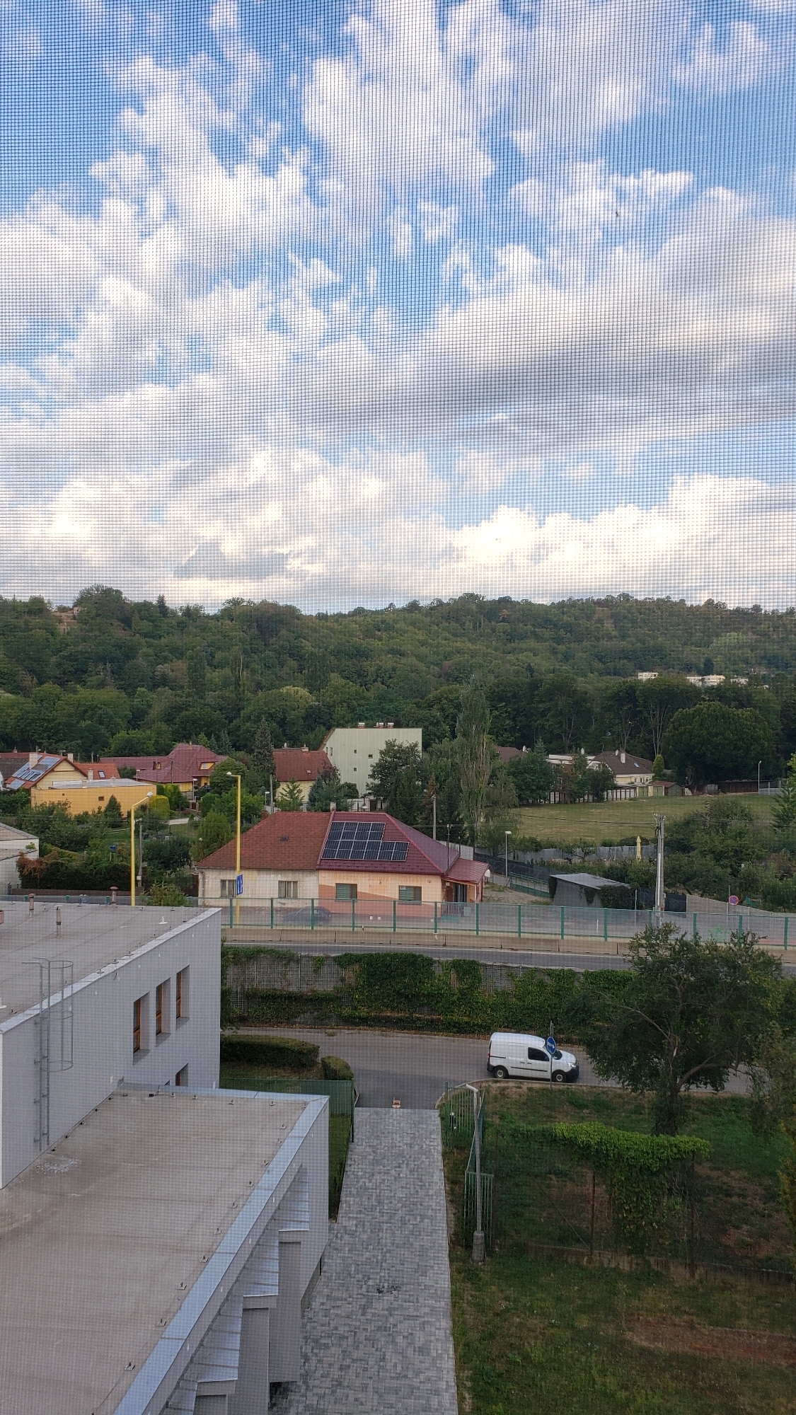 This image depicts a view from a window, possibly from an elevated position in Prešov, Slovakia. The foreground shows the corner of a modern building with a grey, flat roof and a section of exterior wall. A walkway made of grey interlocking pavers leads away from the building. Beyond this, a street is visible, with a white van parked on its right side. Further back, a residential area with several houses can be seen. One house prominently features a red roof with solar panels. Behind the houses, a densely wooded hillside forms the backdrop. The sky above is a mix of blue and white clouds, suggesting daytime. The scene captures a typical suburban or semi-urban landscape.