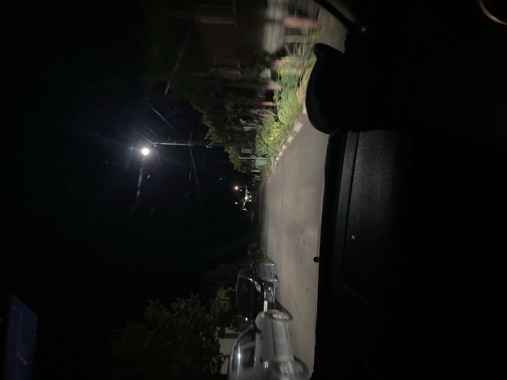 The image is a night-time, interior view from a vehicle driving through the city of Yasen, Bulgaria. The road is paved and appears to be a residential street. On the left side of the road, a silver car is parked, and further down, a black minivan is also parked. Trees and vegetation line the right side of the road, with some houses visible in the background. A street light illuminates the scene. The dashboard of the vehicle is visible in the foreground. The overall atmosphere is dark and quiet.