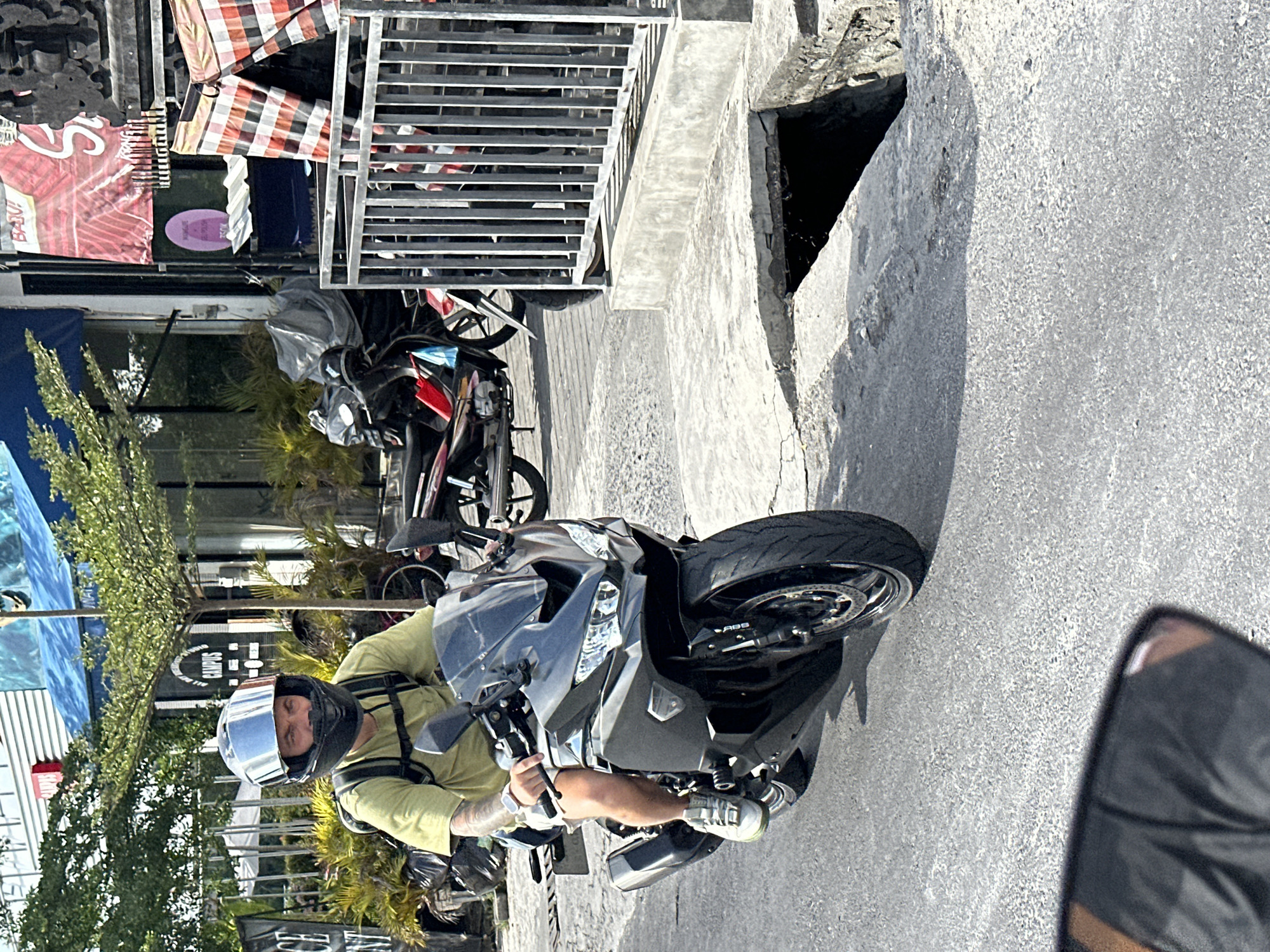 In North Kuta, Indonesia, a person wearing a reflective silver helmet and a light green t-shirt is riding a dark gray sport motorcycle on a paved road. The rider has tattoos on their left arm and is wearing white sneakers. Behind the motorcycle, a black and red motorcycle is parked near a metal fence and some buildings with banners. The scene includes tropical foliage and a concrete structure with an open drain.