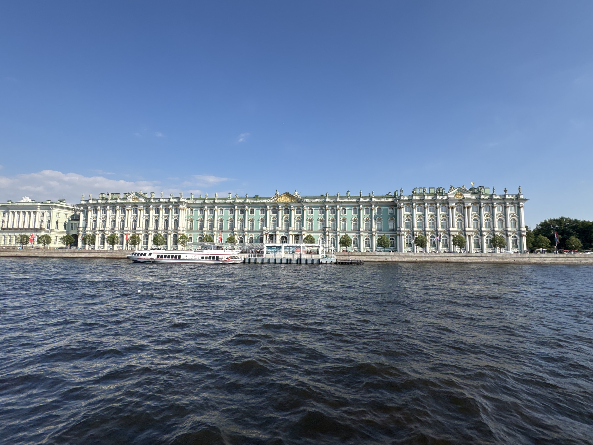 The image shows a wide view of the Neva River in Saint Petersburg, Russia, with a grand, long building situated along its embankment. The building, identified as the Winter Palace, is a prominent architectural landmark, featuring a striking green facade with white decorative elements and numerous windows. The palace is adorned with statues and intricate detailing along its roofline. In front of the palace, a white passenger boat with red trim is docked, and another similar boat is moving on the river. The water of the Neva River is dark blue with visible ripples. The sky above is a clear, bright blue with a few wispy clouds. The scene conveys a sense of grandeur and historical significance, characteristic of the city's imperial architecture.