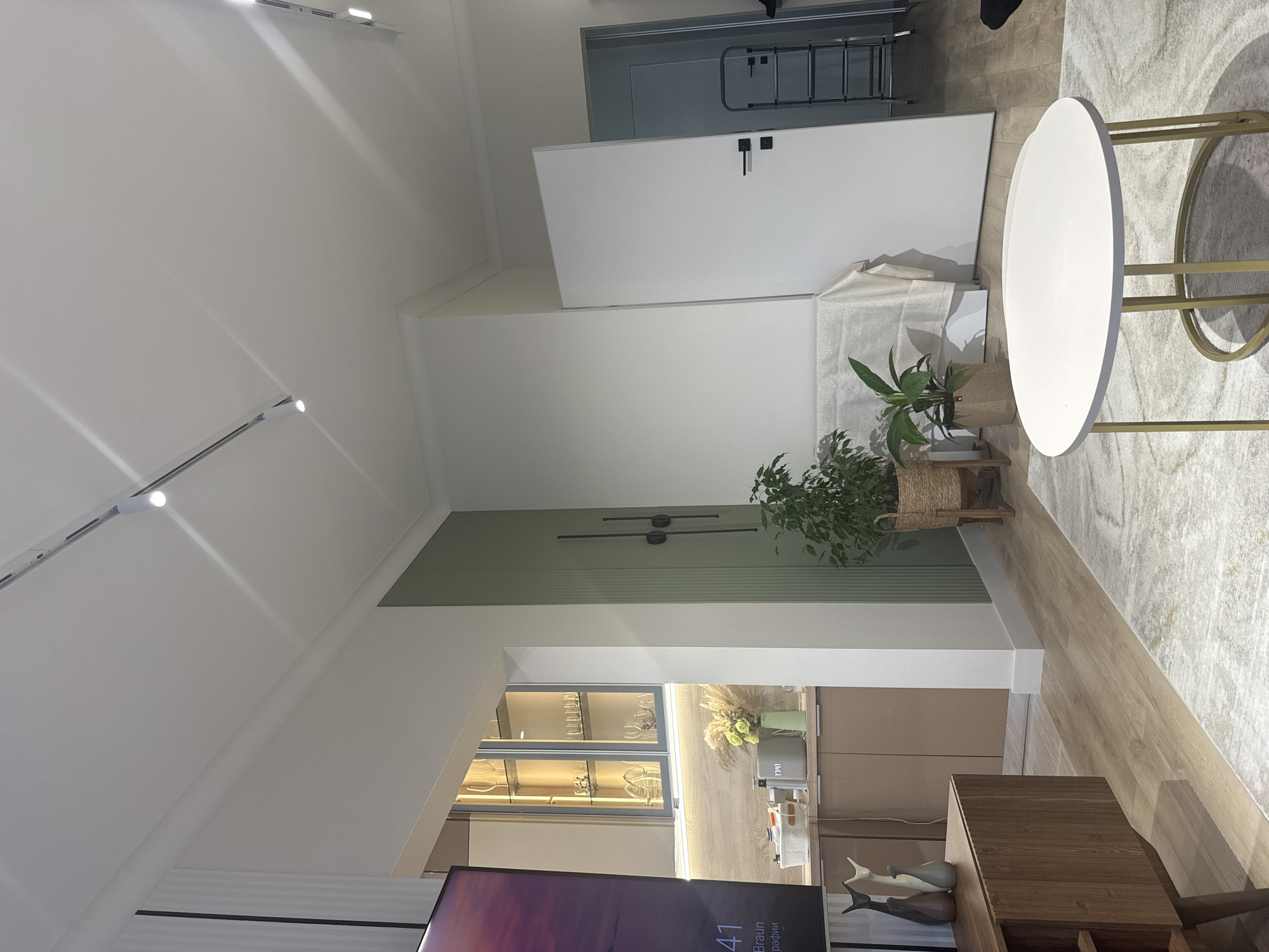 A slightly tilted indoor shot shows a contemporary interior design. A green accent wall features two horizontal black lines. Below is a cabinet with glass doors filled with glasses, and beige cabinets. Potted plants rest on a small wooden stool. A white round table on golden legs stands on a patterned rug. A white door with a black handle is visible, as well as a metal ladder in the background. A television screen occupies the bottom of the frame. The image includes text "Braun графии" and "12:41" (time), presumably on the TV. There is no indication of people, their interactions or specific activities. Location details (Nizhnevartovsk, Russia) are unsupported by the image.