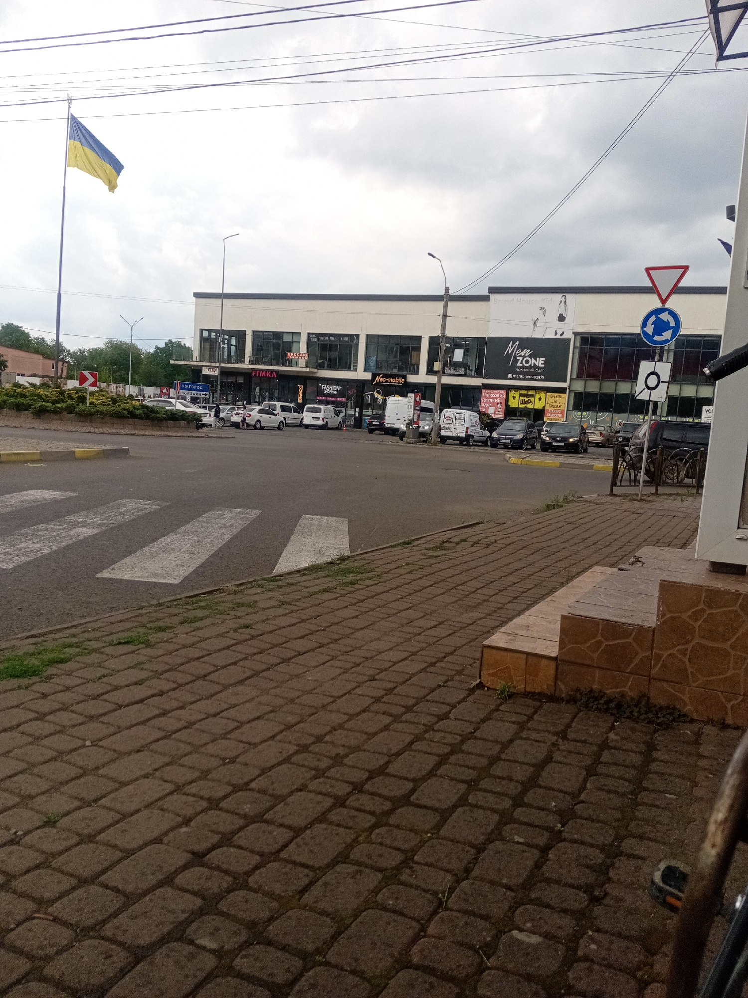 The image shows a street scene in Tiachiv, Ukraine, on a cloudy day. In the foreground, a pedestrian crosswalk is visible, made of white stripes on asphalt. The sidewalk is paved with dark bricks. To the left of the crosswalk is a small, circular island with green bushes and a red and white directional sign. In the background, a multi-story building with a modern facade houses several businesses, including "Men ZONE" and "FIMKA." A Ukrainian flag flies from a tall pole to the left of the building. Several cars are parked and driving on the street. The overall impression is a typical urban environment with commercial activity.
