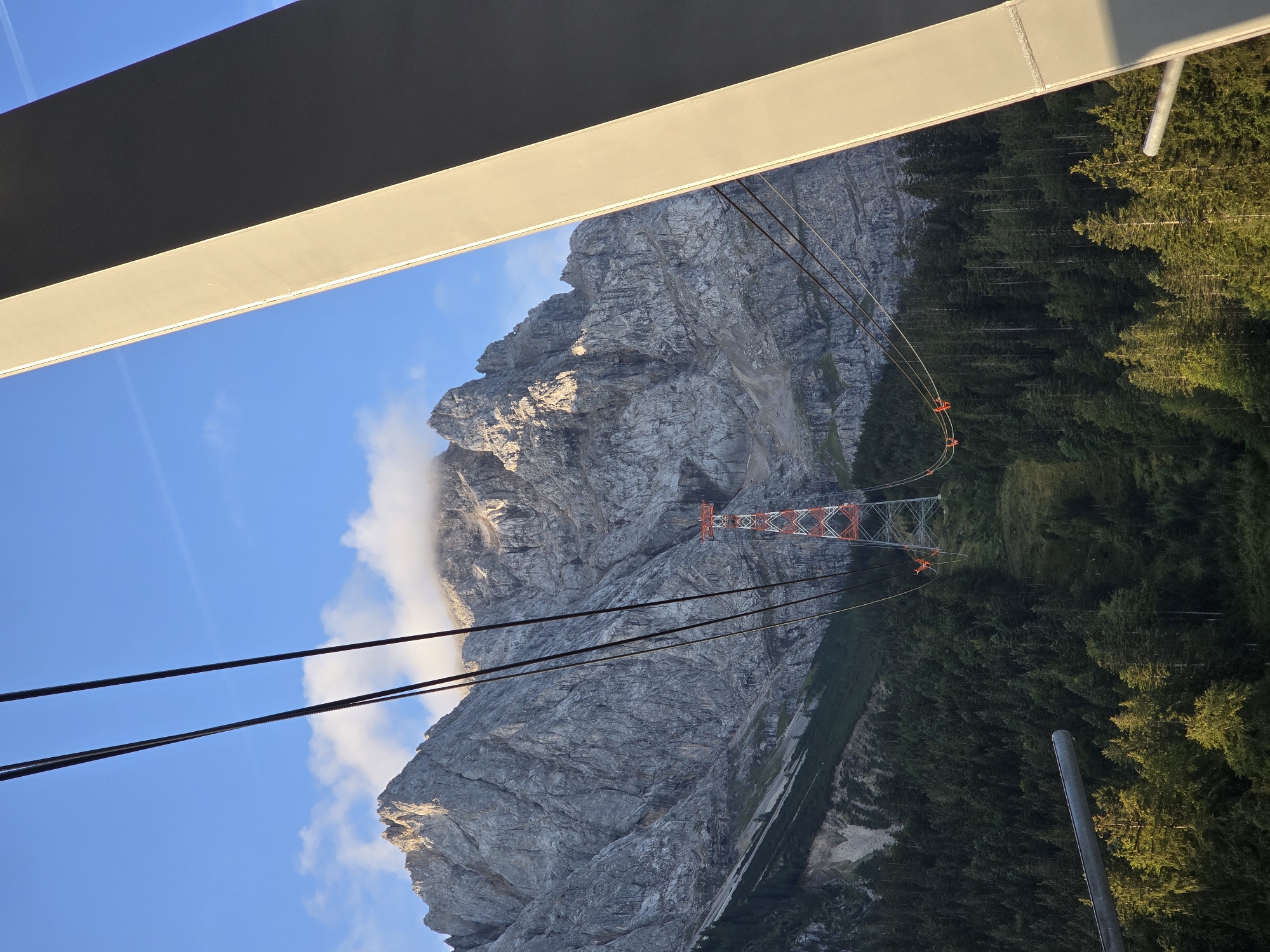 The image, captured from a gondola or cable car ascending the Zugspitze mountain in Grainau, Germany, presents a dramatic mountainous landscape. In the foreground, a large, light-colored, metallic support structure dominates the right side of the frame, casting a shadow. Several thick cables, likely part of the cable car system, stretch diagonally across the sky and towards the mountain.
The middle ground features a towering, rugged mountain, its rocky slopes textured and showing signs of erosion. A distinct red and white pylon, a support for the cable car, stands prominently on the mountainside, with its cables clearly visible. Below the pylon, the mountain transitions into a dense, dark green forest, primarily composed of coniferous trees. Patches of lighter green indicate grassy slopes amidst the trees. A few small, bright orange figures, presumably people, are visible on the cable car system near the pylon, appearing to be maintenance workers or passengers.
The background is a clear blue sky, dotted with a few wispy white clouds, one of which is partially obscuring the peak of the main mountain. The sunlight illuminates the upper parts of the mountain and the forest, creating a play of light and shadow. The overall scene evokes a sense of grandeur and elevation, typical of a high mountain resort.