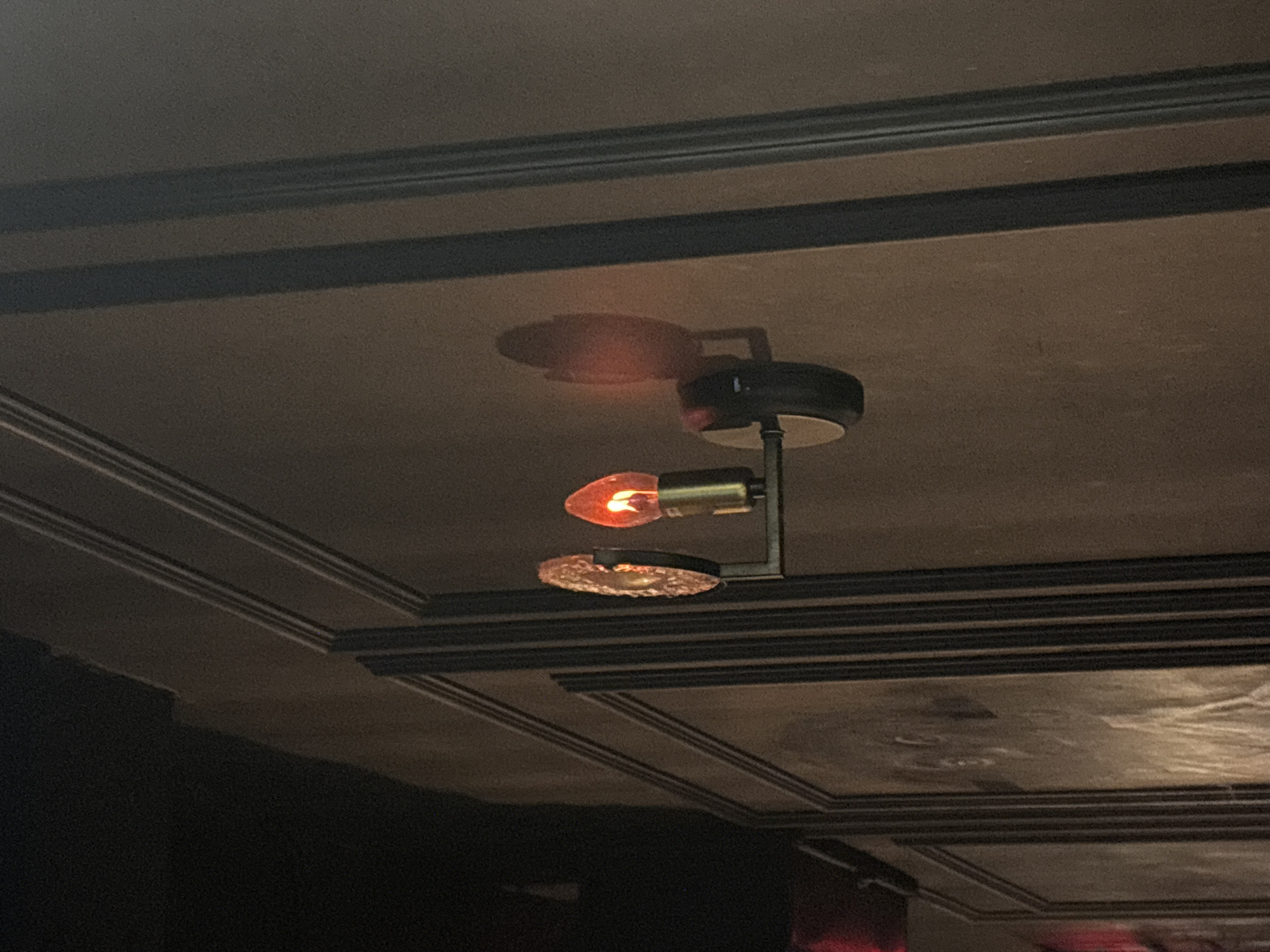 A close-up, eye-level shot captures a dimly lit wall with ornate paneling and a vintage-style light fixture. The light fixture, made of dark metal, holds two decorative elements: a textured, circular glass or metal disc to the left and an illuminated, red, teardrop-shaped bulb to the right. The bulb emits a warm, orange glow, casting a soft shadow on the wall behind it. The wall itself is textured and appears to be a muted brown or grey color, divided into rectangular panels by vertical moldings. The ceiling above is dark and out of focus. The image does not provide any information about the city of Kazan, Russia, or any specific people, actions, or interactions.