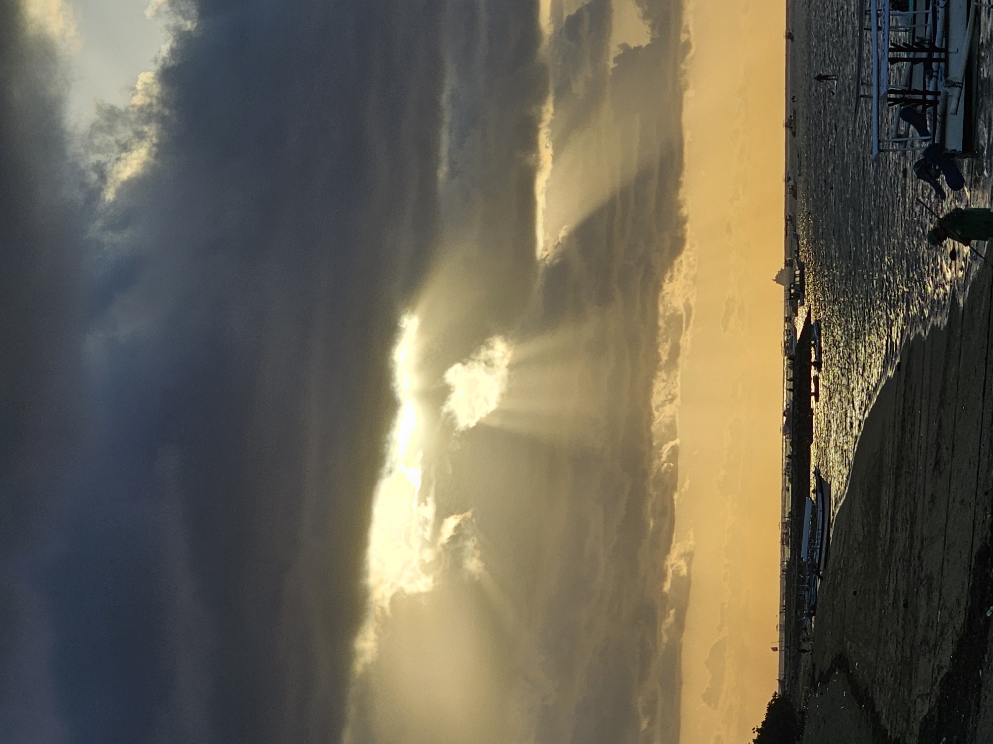 The image depicts a coastal scene, likely in Denpasar, Indonesia, during what appears to be a dramatic sunset or sunrise. Dark, ominous clouds dominate the upper portion of the frame, with shafts of golden light breaking through, creating a spotlight effect on the water. The sea is choppy, reflecting the light in a shimmering pattern.
On the shore in the foreground, a dark, sandy beach is visible, with some debris scattered along its edge. Several boats are moored or anchored in the shallow water. Some of these appear to be traditional outrigger boats, while others are larger vessels, possibly tour boats.
In the mid-ground, a line of boats stretches along the horizon, and further out, a larger ship can be seen. There are scattered figures on the beach and in the water, some appearing to be walking or engaged in activities. One person is visible in the lower right corner, near a boat, and another is on a paddleboard in the distance. The overall atmosphere is one of natural beauty and a sense of transition, perhaps due to the weather or time of day.