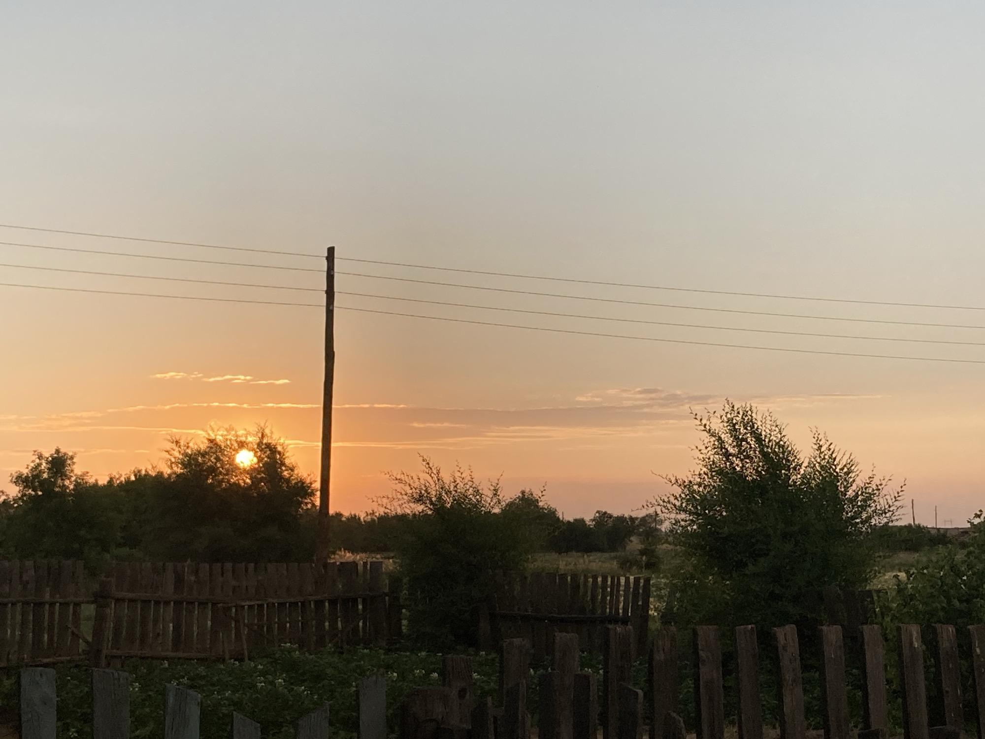The image depicts a rural scene at sunset in Rakity, Russia. The sun is low on the horizon, casting a warm, orange glow across the sky and illuminating the clouds with soft, pastel hues. A rustic wooden fence runs horizontally across the foreground, with several wooden posts scattered in front of it. Behind the fence, lush green bushes and trees create a dense backdrop. A single wooden utility pole stands vertically on the right side of the image, with several horizontal wires stretching across the sky. The overall mood is serene and tranquil, evoking the peaceful atmosphere of a rural evening. No people are visible in the image.