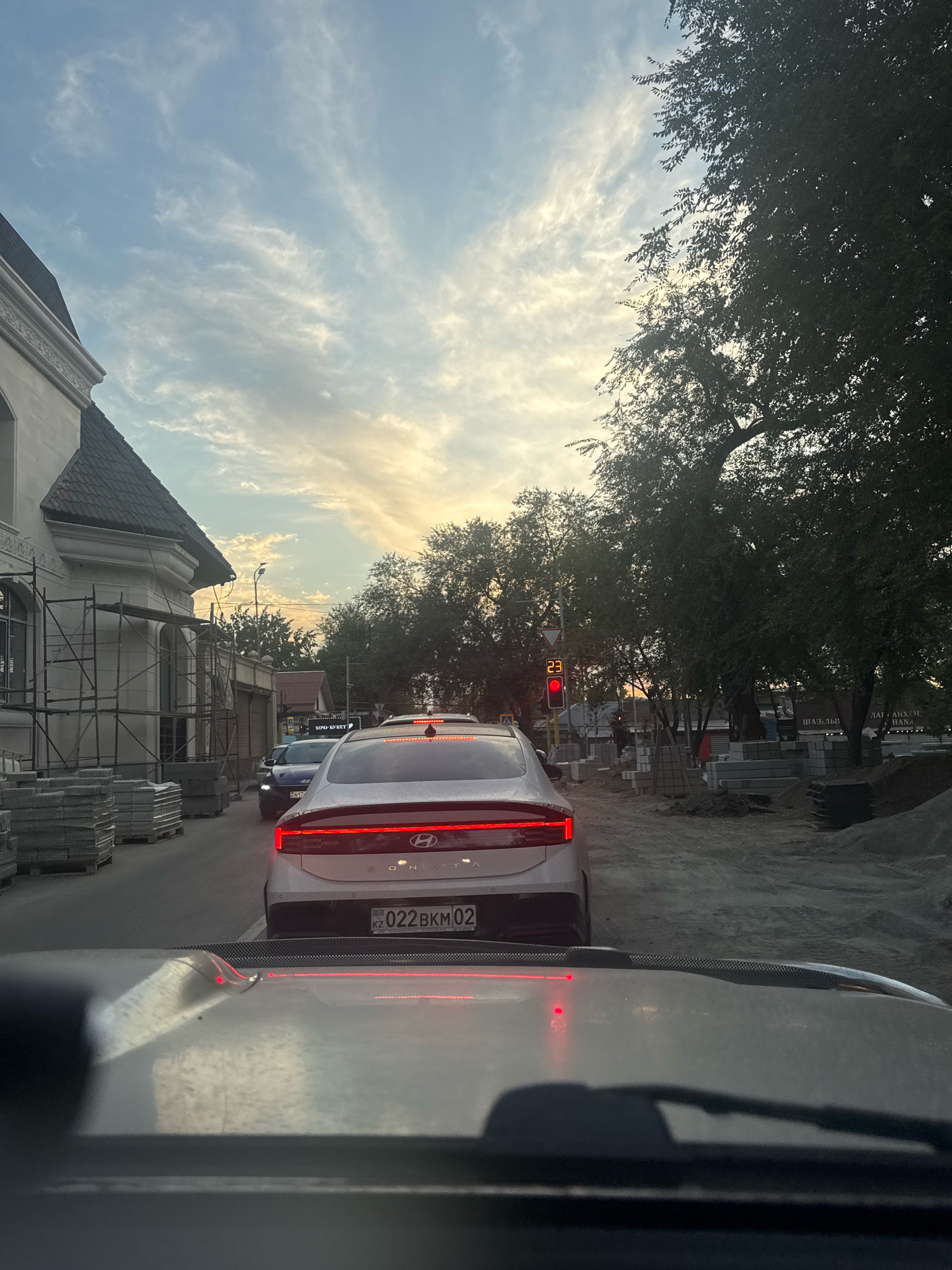 The image depicts a street scene in Almaty, Kazakhstan, during daylight hours. The perspective is from inside a vehicle, looking forward. Traffic is stopped, indicated by the red traffic light illuminated with the number "23". On the left side of the road, a building under construction is visible, with scaffolding and stacks of building materials, possibly pavers. Several cars are in traffic, including a white Hyundai Sonata with a Kazakhstan license plate that reads "KZ 022 BKM 02". On the right side, trees line the street, and more construction materials, including piles of gravel and stacks of pavers, are present. The sky is partly cloudy with a warm glow, suggesting either sunrise or sunset.