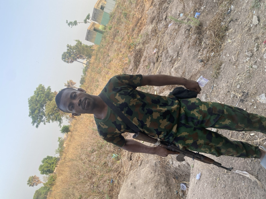 A man in camouflage fatigues poses outdoors, holding an AK-47-style rifle. He has a black bag slung across his body. The backdrop features a rural landscape with dry, grassy terrain, scattered trees, and distant buildings. The sky is hazy, suggesting possible dust or atmospheric conditions. The man appears to be in a field or clearing, with debris or litter visible on the ground.