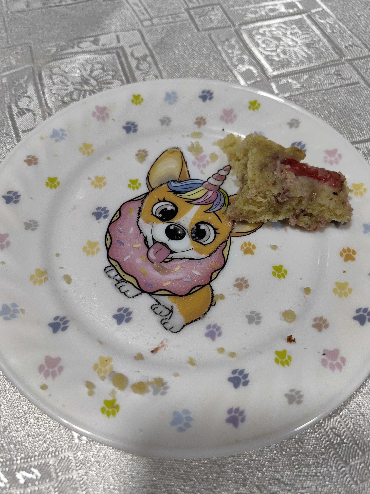 A plate with a drawing of a corgi wearing a donut and a unicorn horn is shown. There is a piece of cake with red jam on the plate, next to the corgi drawing. The plate is white with colorful paw prints. The plate is placed on a table with a patterned tablecloth. The image is taken from a top-down perspective. No people are visible in the image. The location is unspecified.