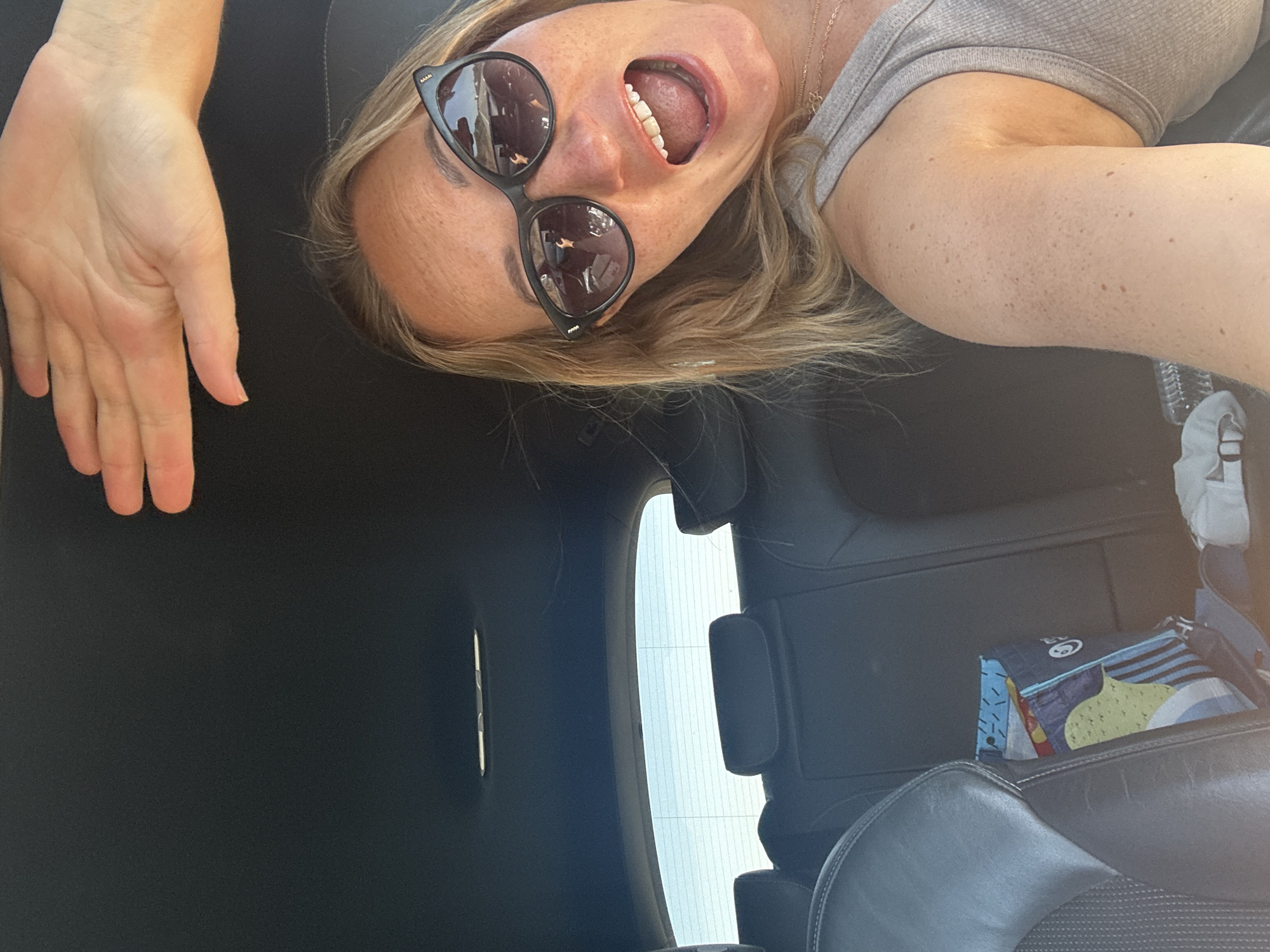 A woman is in the driver's seat of a car, with her right arm raised above her head, obscuring part of the image. She is wearing sunglasses and has her mouth open in a yell or laugh. The interior of the car is visible, including black leather seats and the headliner. A colorful tote bag is on the passenger seat. The background outside the car is bright and appears to be a sunny day. The scene is set in Olhão, Portugal.