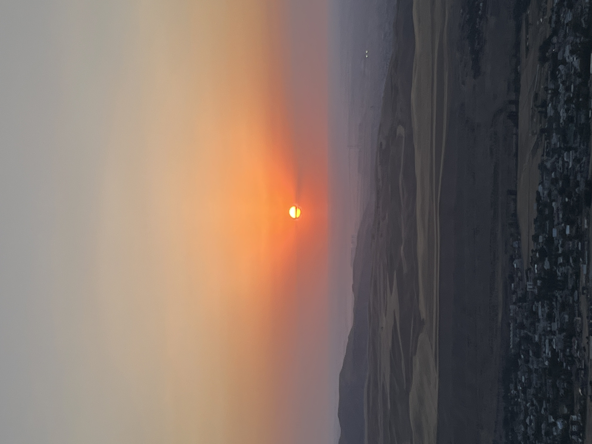 A high-angle shot captures a sunset over a vast, arid landscape with a town in the foreground. The sun, a bright orange orb with a smaller halo, is positioned centrally in the frame, casting a warm glow across the sky, which transitions from a pale yellow at the horizon to a soft grey at the top. Below the sky, rolling hills and distant mountains are silhouetted against the fading light. The terrain appears dry and barren, with visible undulations in the land. In the lower portion of the image, a town is spread out, characterized by densely packed buildings with dark rooftops. The urban area appears to be nestled within the natural landscape, with roads and scattered vegetation visible between the structures. The overall impression is one of a remote, underdeveloped settlement bathed in the warm hues of a sunset. The exact location is not discernible from the image alone; however, the prompt specifies it to be the city of Besh Kungei, Kyrgyzstan.