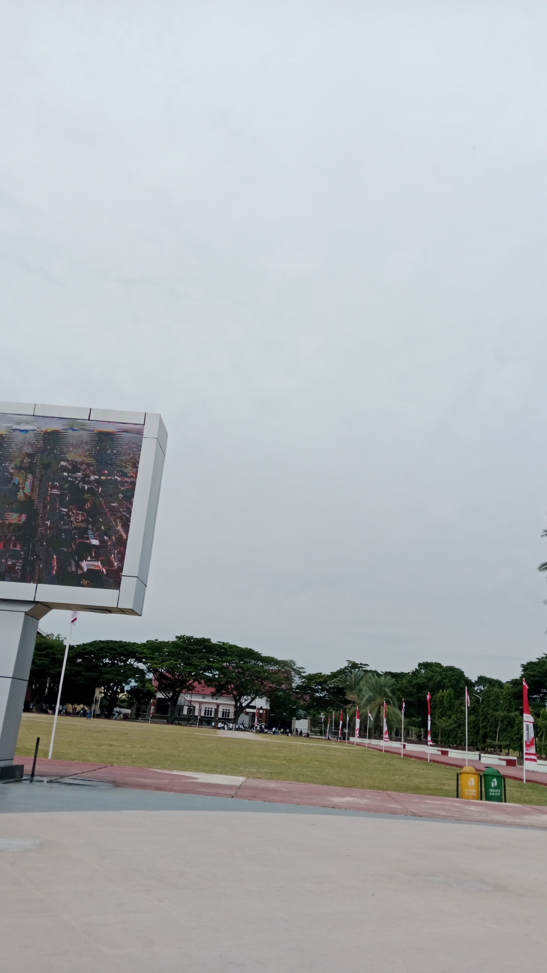 The image depicts a wide, open grassy field in what appears to be a public space, possibly a park or a city square, in Lhokseumawe, Indonesia. In the foreground, a large, modern electronic billboard is visible on the left side, displaying an aerial view of a city. The billboard is mounted on a white, angular structure. The field itself is vast and green, with a red and white checkered border along one edge.
In the mid-ground, a line of trees with lush green foliage forms a natural boundary. Behind the trees, a red-roofed building with white accents is visible. Several people are scattered across the field, some walking and others appearing to be gathered in groups, suggesting a public event or casual activity. A series of Indonesian flags on tall poles are planted along the edge of the field, with one flag visible in the far left of the image.
Further to the right, two trash bins are placed near the red and white border; one is yellow and the other is green. The sky is overcast and a pale gray, indicating a cloudy day. The overall impression is of a well-maintained urban open space, likely a significant gathering point in Lhokseumawe.