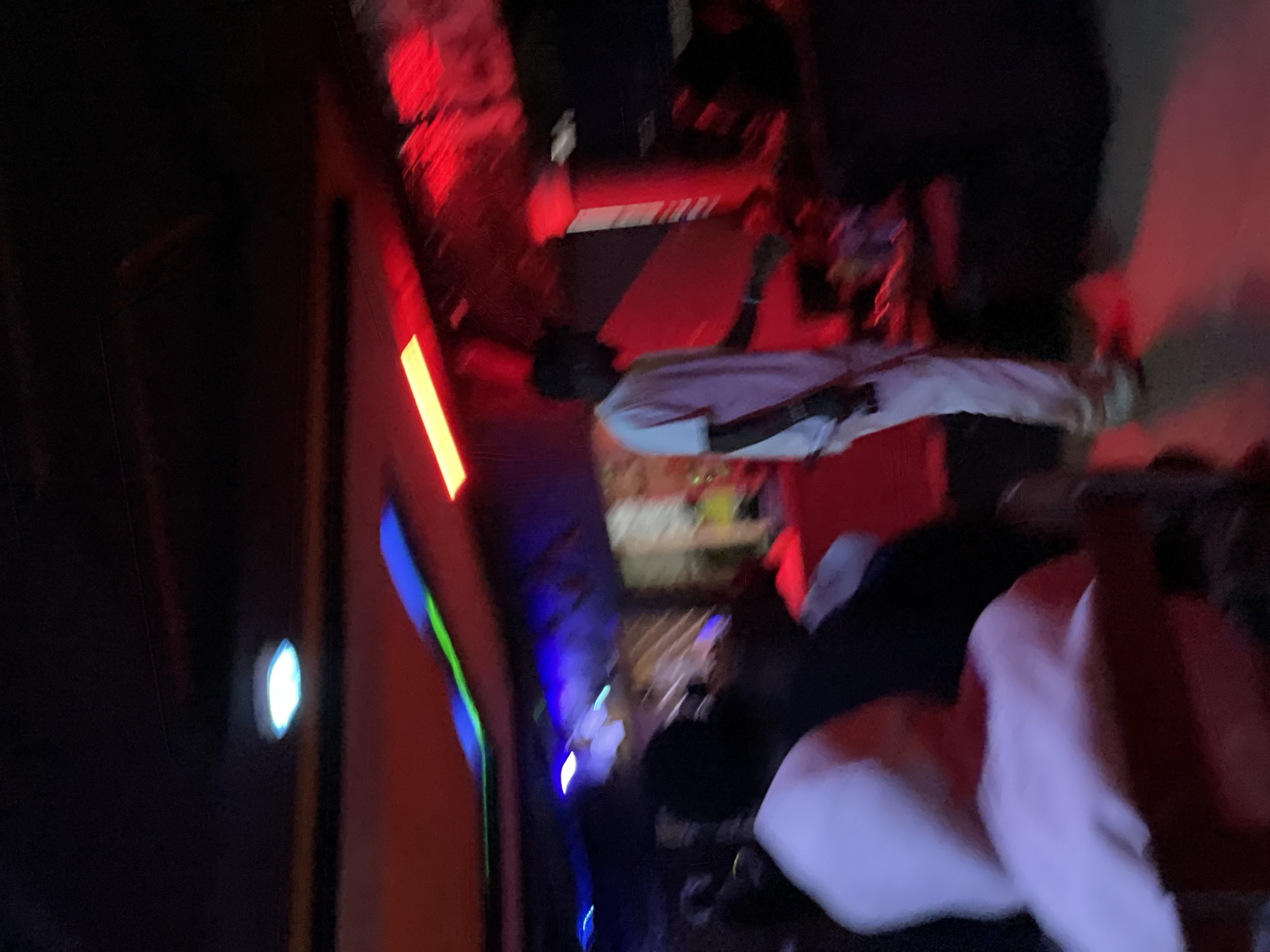 The image depicts an indoor scene with a person standing in the center. The environment appears to be a dimly lit establishment, possibly a club or lounge, with vibrant red and blue lighting. The person in the center is wearing light-colored clothing, including a t-shirt and pants, and is facing away from the camera. Other individuals are present in the background, but their features are obscured by low light and motion blur. The ceiling features a multi-layered design with integrated lighting. The flooring is dark and reflects some of the colored lights. The scene suggests an active social gathering, potentially at night. The context of Durumi, Nigeria, suggests this scene is likely within an urban entertainment venue in that specific location.