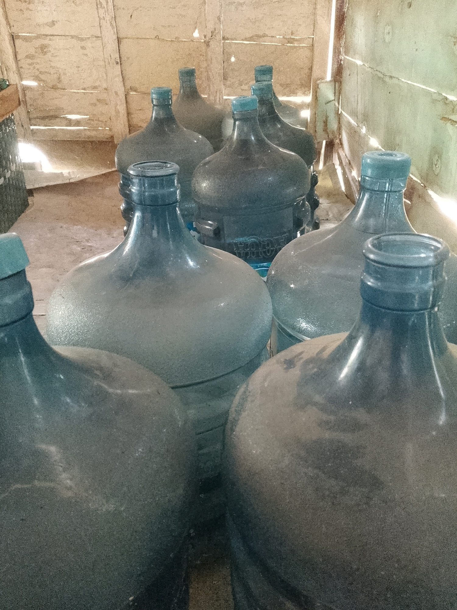 In the city of Wadang, Indonesia, a collection of dusty, blue, five-gallon water jugs is arranged in an interior space. The jugs, some filled and some empty, are clustered together, with their blue caps visible. The surrounding environment consists of a wooden wall constructed from vertical planks, with light filtering through gaps between them, creating dramatic shadows and highlighting the dust on the jugs. The floor appears to be made of concrete. The scene is dimly lit, with natural light sources creating a somber atmosphere.
