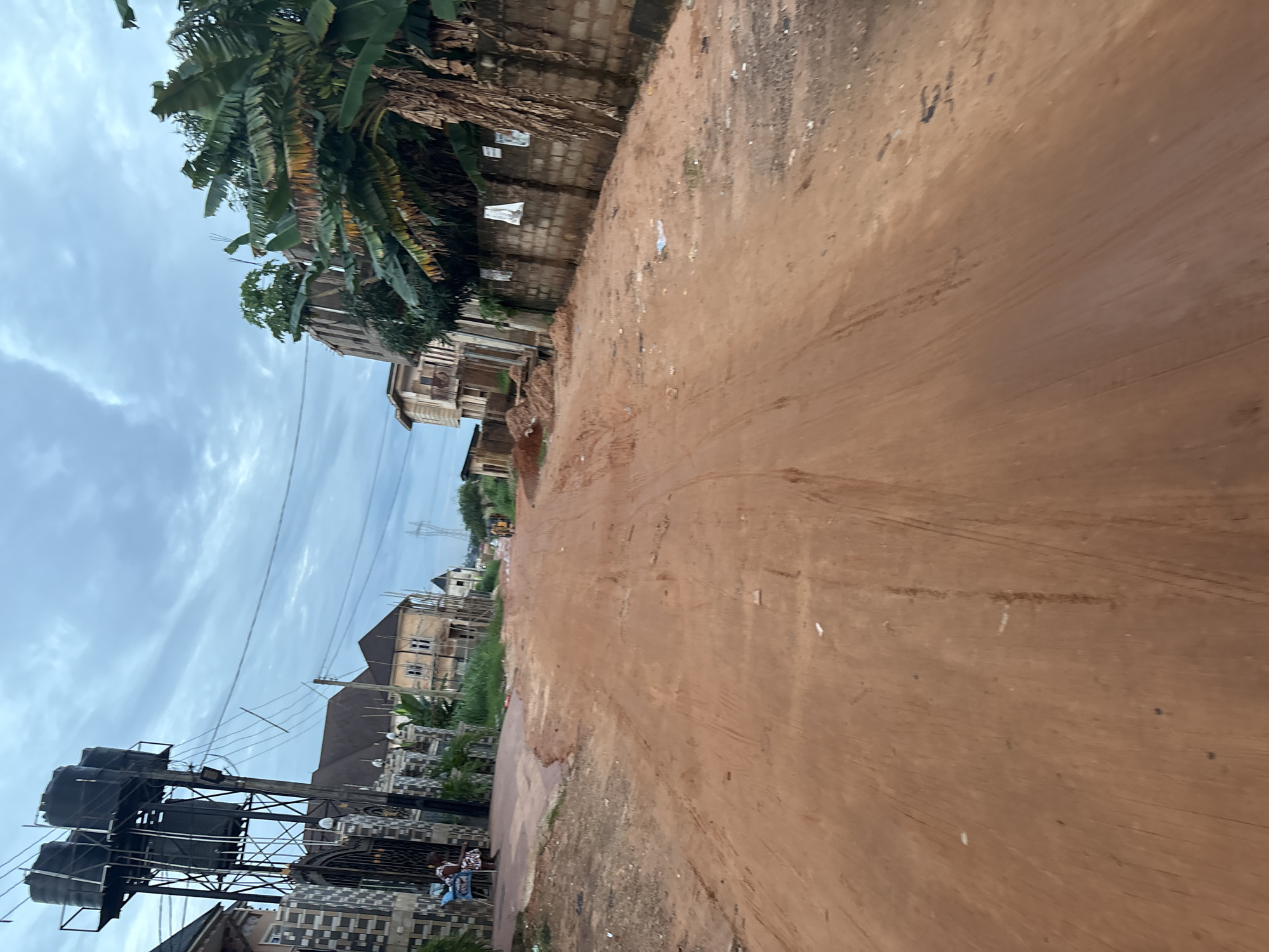 The image depicts a dirt road in Obosi, Nigeria, likely during daylight under an overcast sky. The road is unpaved and characterized by red soil with visible tire tracks and some scattered debris, including small pieces of white paper. On the left side of the road, a tall metal structure supports multiple large black water tanks. Adjacent to this structure is a gated entrance to a property with decorative fencing. Further down the road on the left, there are several multi-story buildings, some appearing to be under construction with exposed rebar. On the right side of the road, a brick wall runs along the property line, partially obscured by lush green banana trees and other foliage. There are posters attached to the wall. In the distance, at the end of the road, two individuals are visible, one appearing to be walking, and a few other buildings and trees can be seen. The overall scene suggests a developing or semi-developed residential area.