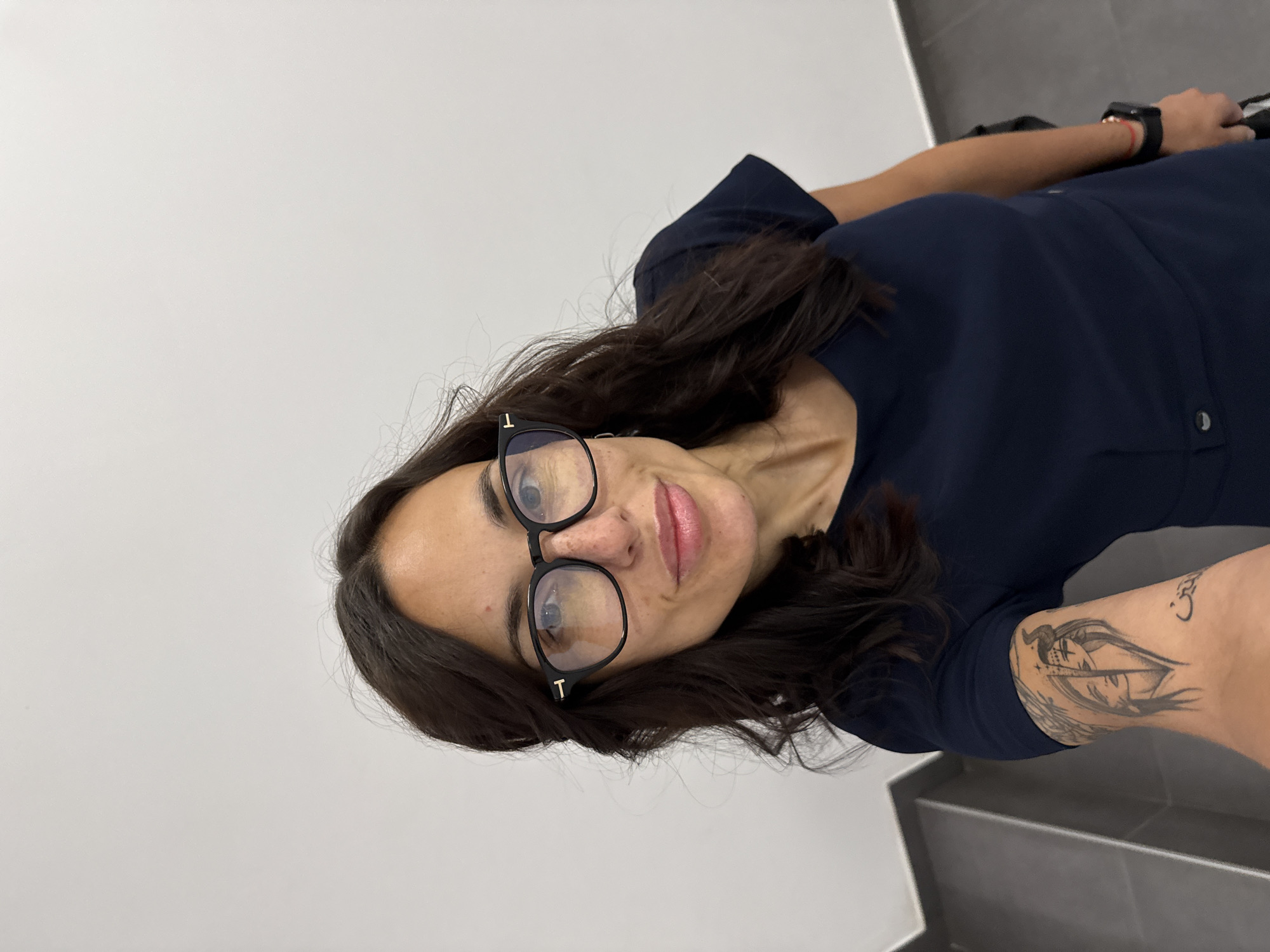 A woman with dark, wavy hair and wearing glasses is seen in a selfie-style photograph. She has a fair complexion and is wearing a dark blue dress or top. Her left arm is visible, featuring a tattoo. The background consists of a plain white wall and a grey, tiled area resembling a bathroom or kitchen surface. The setting does not reveal any information related to Pleven, Bulgaria.
